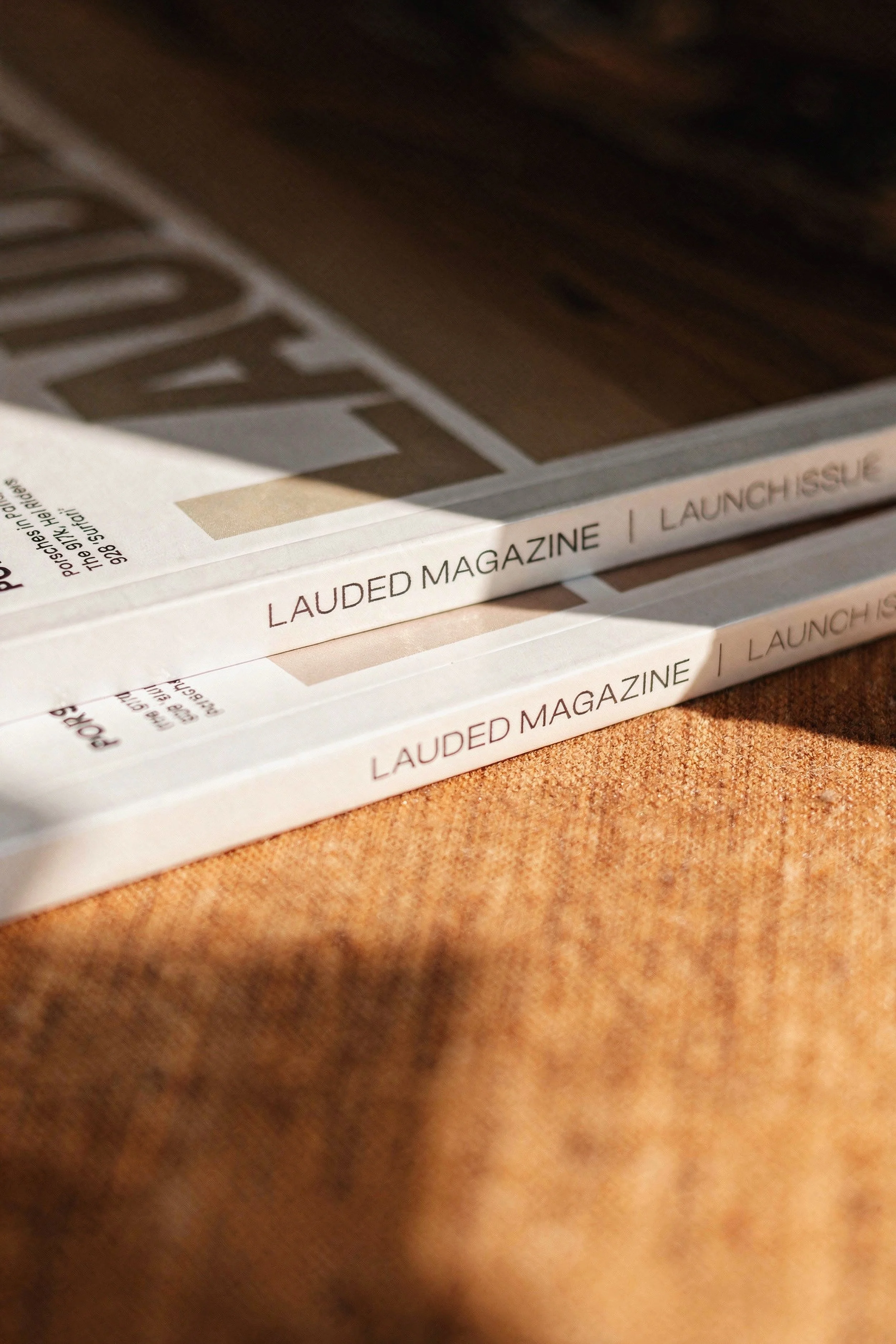 Three copies of Lauded Magazine placed on a textured orange surface, with a wooden background and sunlight casting shadows.