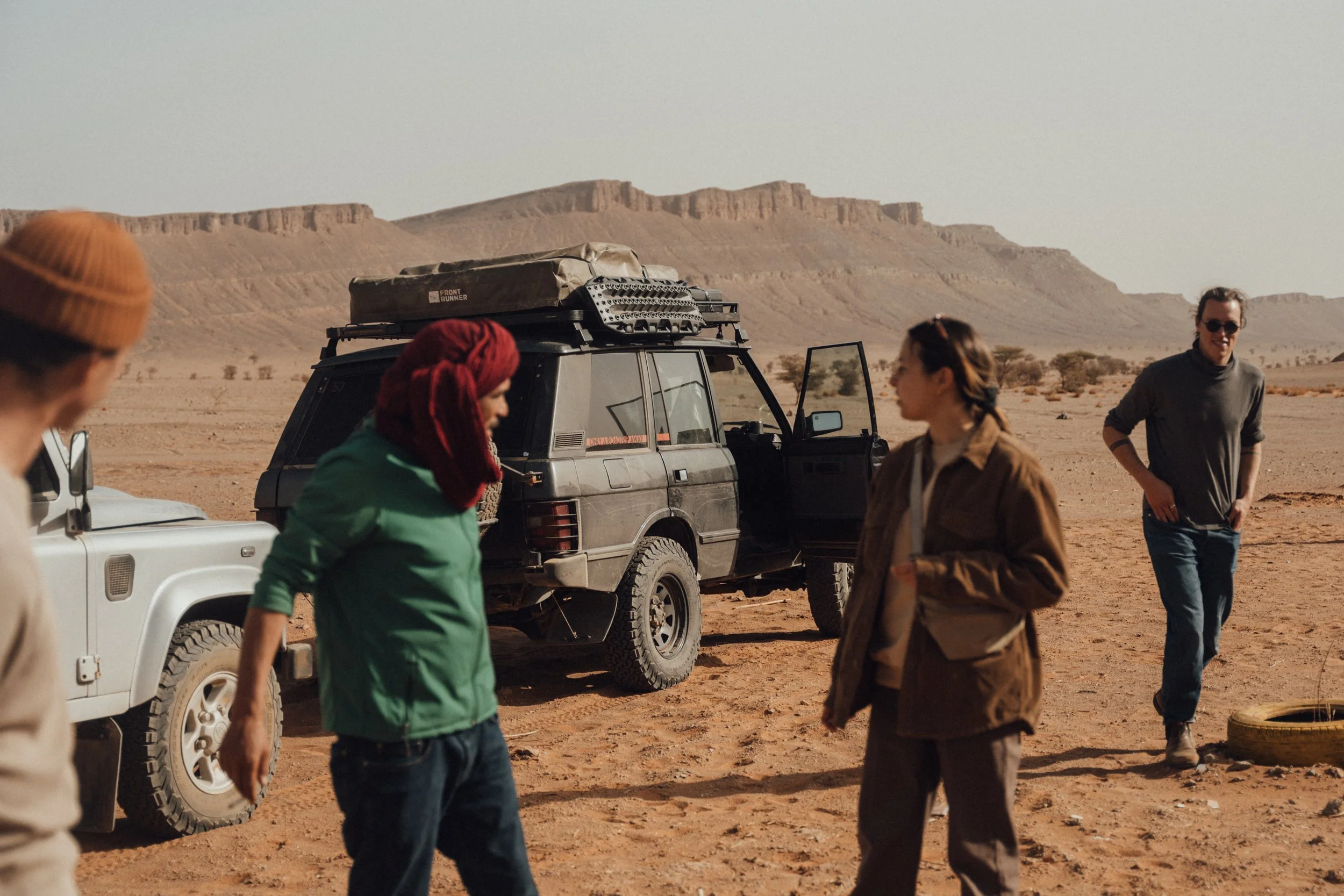 A group of people standing around in a desert landscape with mountains in the background, next to parked off-road vehicles.