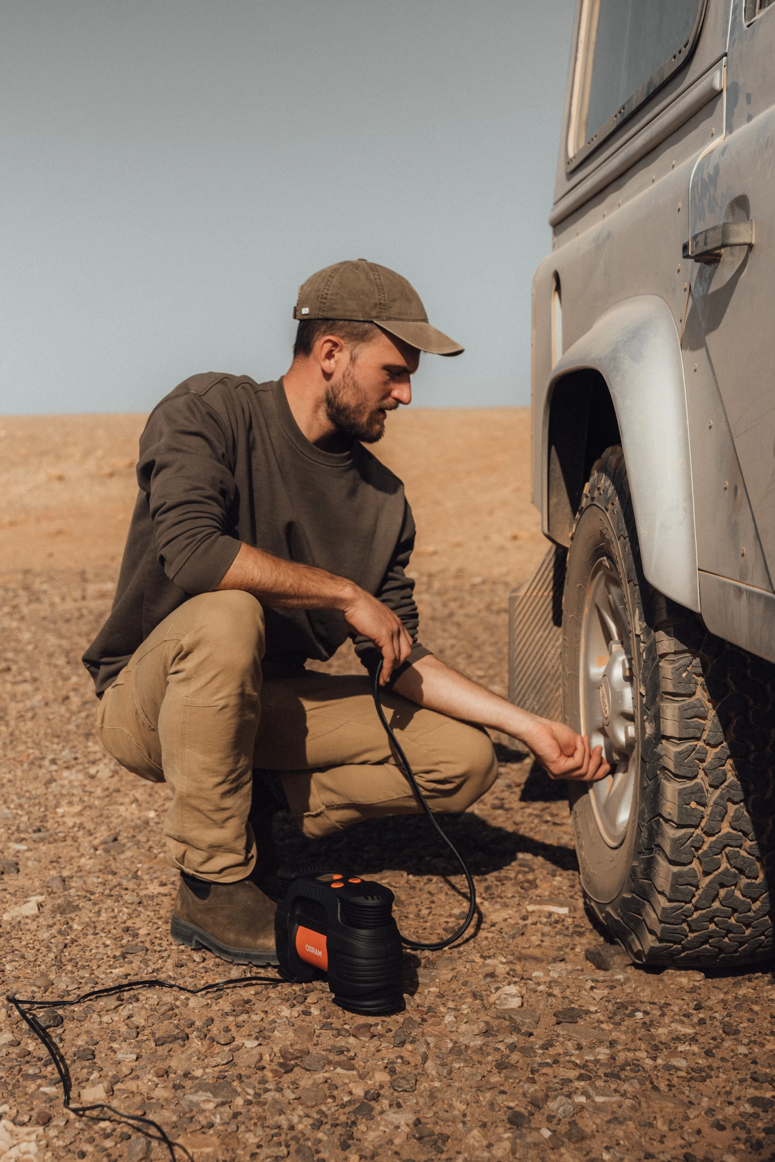 A man crouching beside a truck, using a portable air compressor to inflate the tire in a desert landscape.
