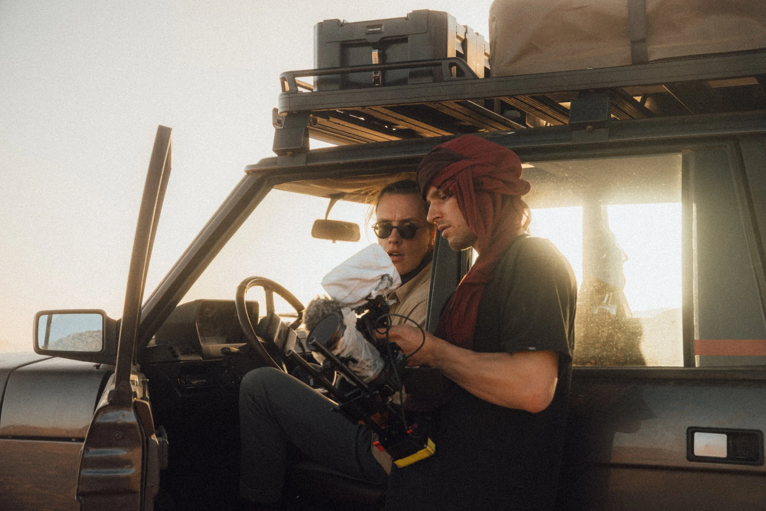 Two people sitting inside a vehicle, with one person holding a robotic device and wearing gloves, and the other person wearing sunglasses and a headscarf, during sunset.