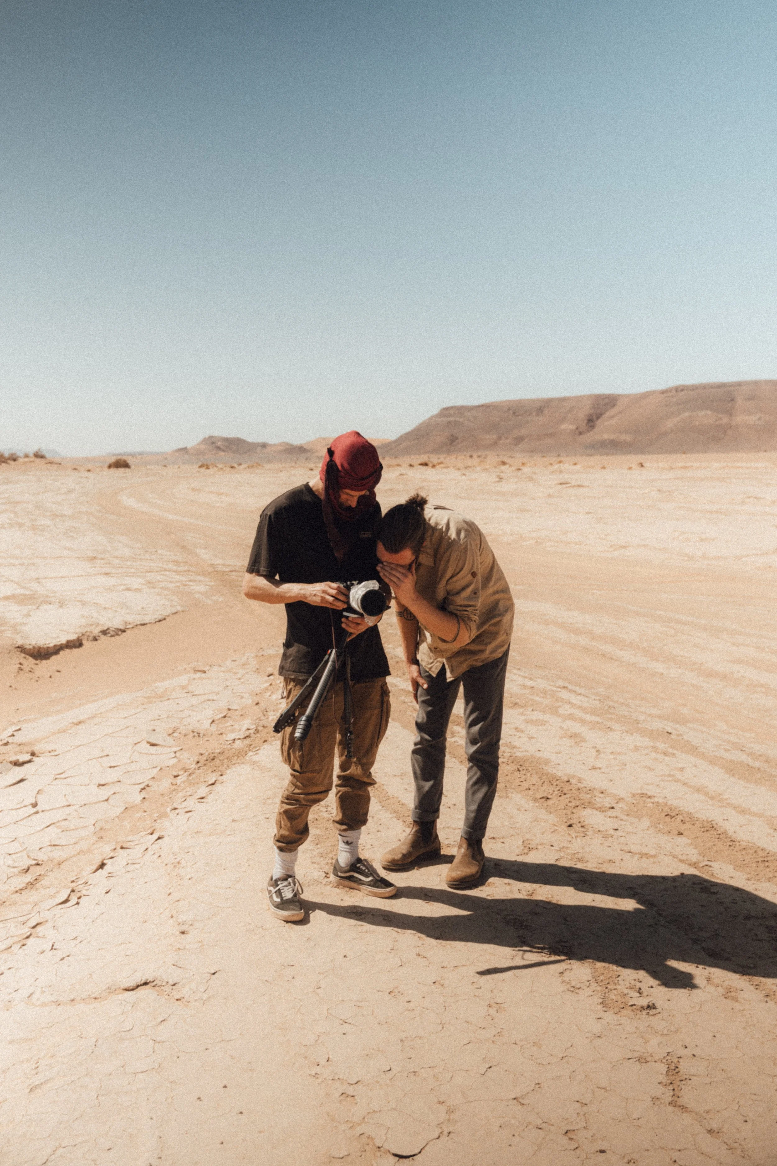 Two people standing in a desert, one holding a camera and the other covering his face with his hand.
