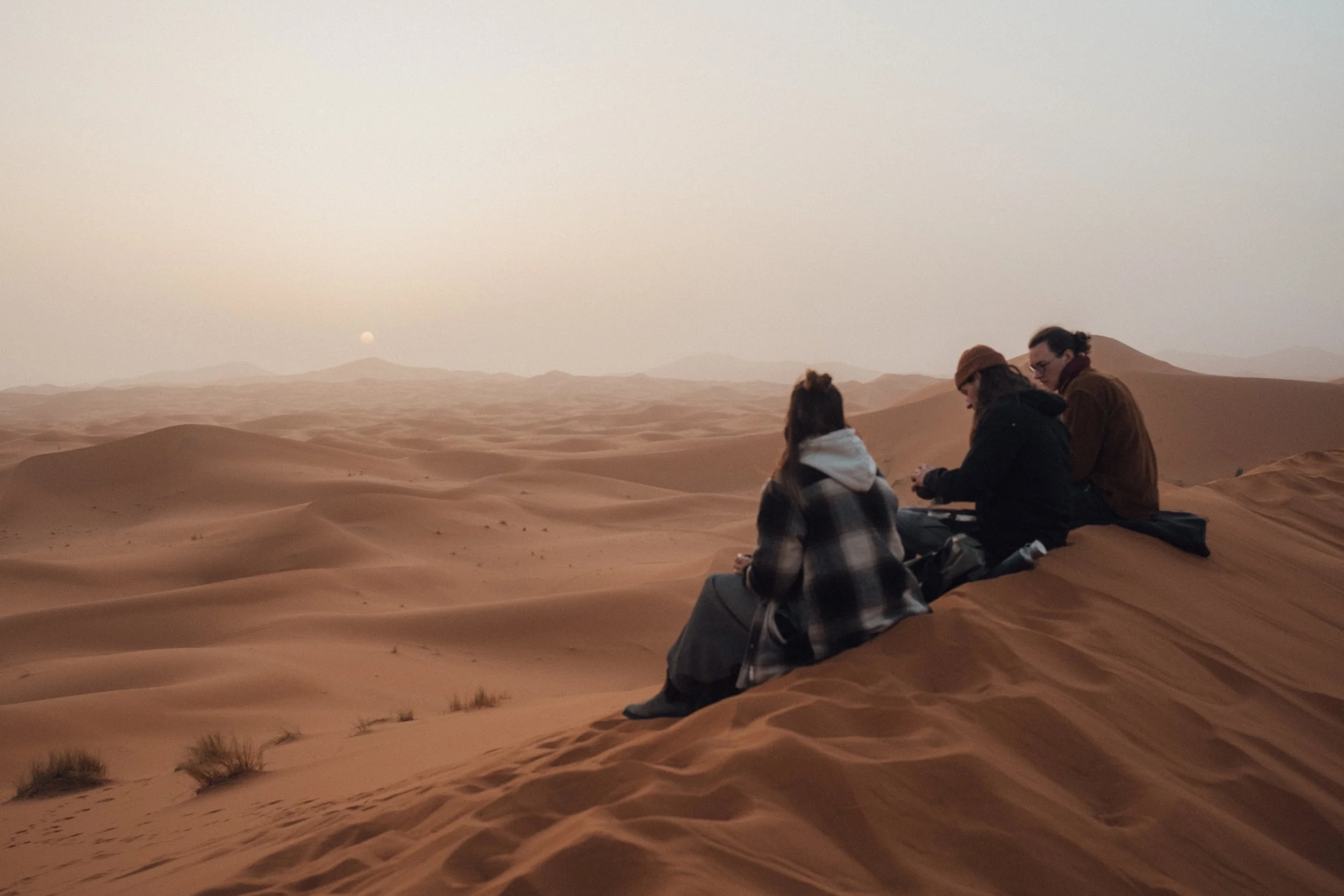 Three people sitting on sand dunes in a desert at sunset or sunrise, with a moon visible in the sky.
