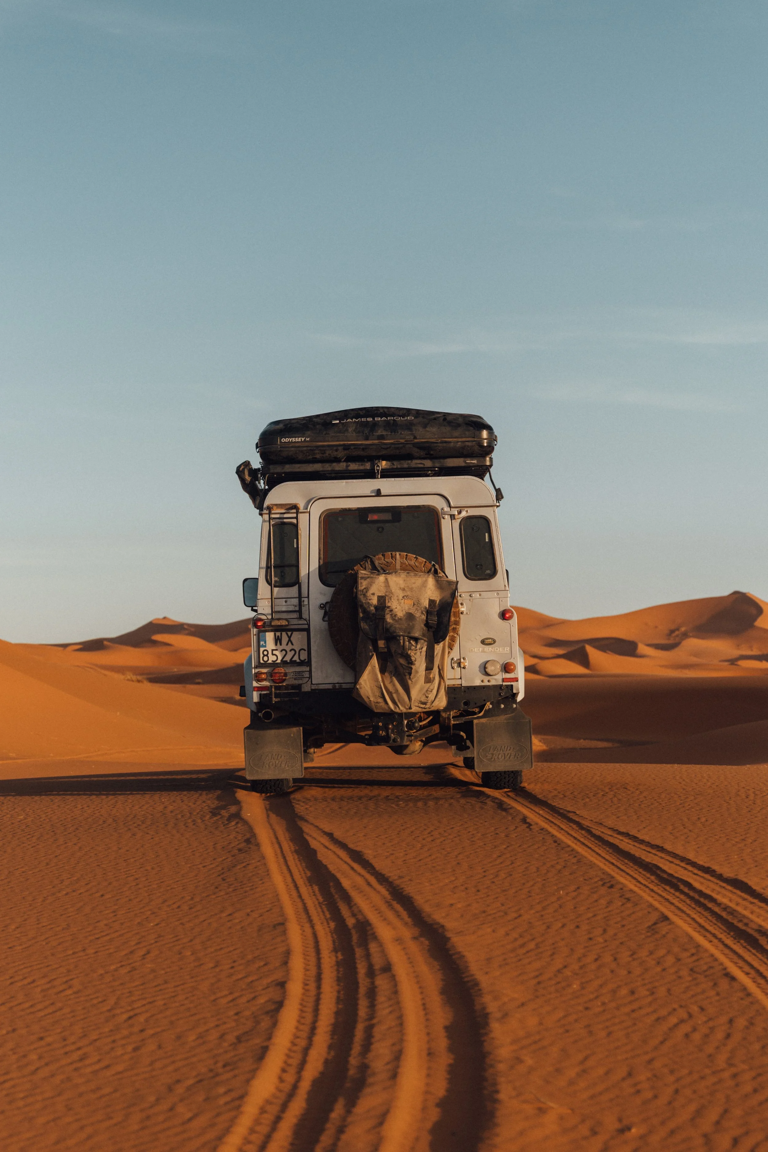A white off-road vehicle driving through a desert with sand dunes, leaving tire tracks behind.