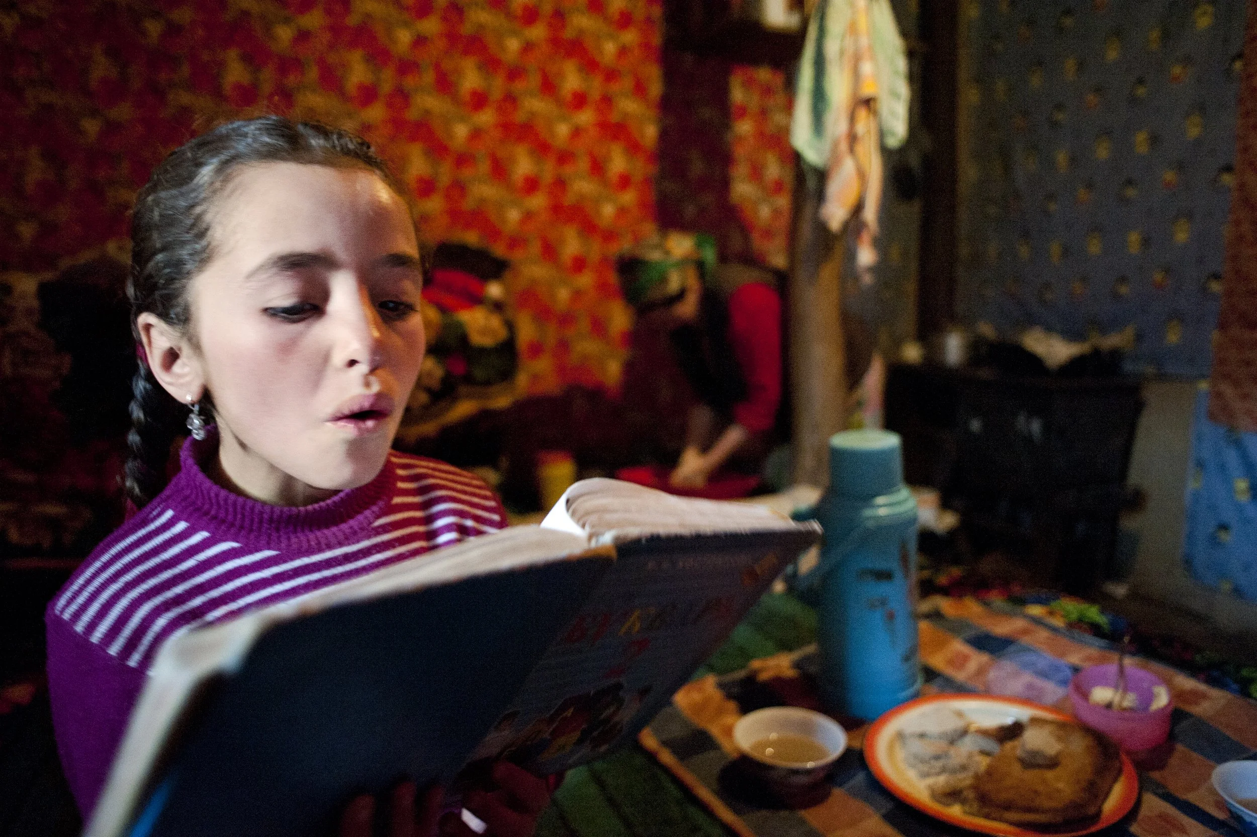 She does her homework after school. Behind, her mum prepares bread for dinner. Lessons in primary school are mixed. Ismailis do not have a madrasa, the Koranic school. At school, she learns Russian and Tajik. She will start learning English at second