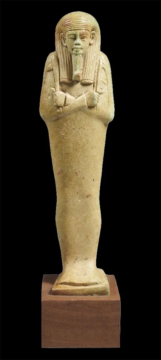 Ancient Egyptian carved stone statue of a pharaoh with crossed arms, wearing a headdress, standing on a rectangular base.