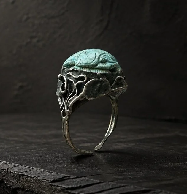 A silver ring with an ornate design and a large greenish stone, shaped like a skull, set against a dark background.