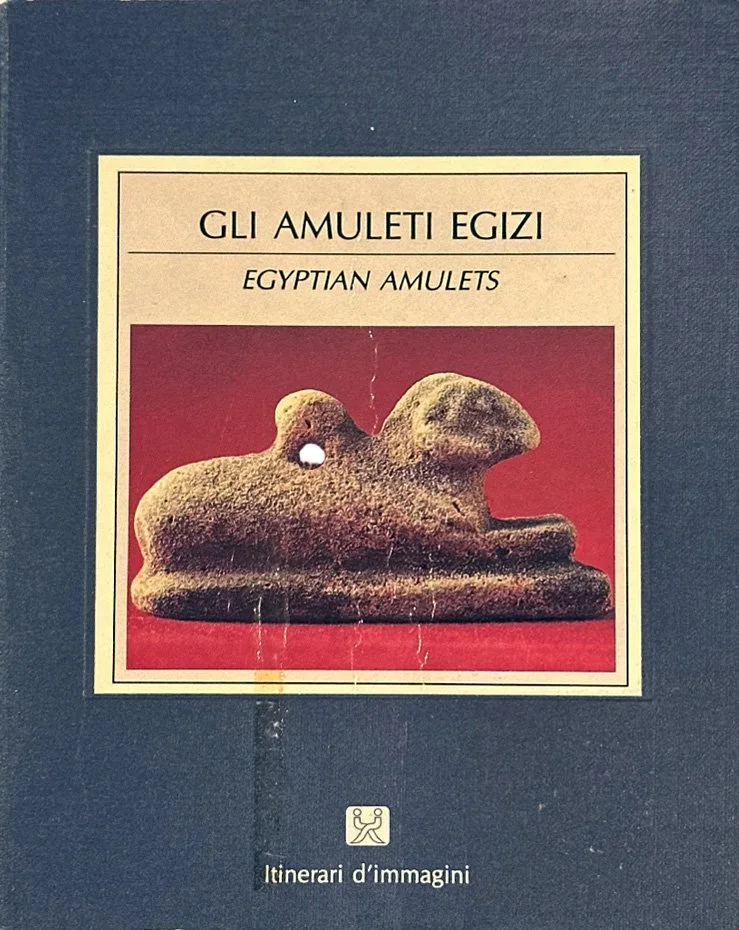 A book cover titled 'Gli Amuleti Egizi' with a picture of an ancient Egyptian amulet shaped like a crouching animal, possibly a dog or a lion, on a red background. The book is published by Itinerari d'immagini.