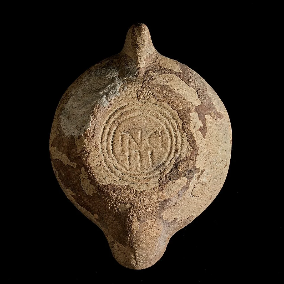 Ancient clay or ceramic object with a circular stamp or imprint marked with the letters 'NCH' on a black background.