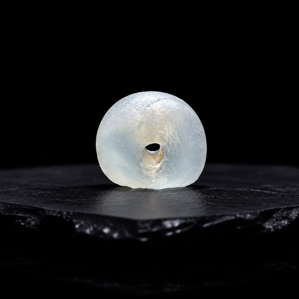 A close-up of a white sea glass bead with a central hole, placed on a black surface against a black background.