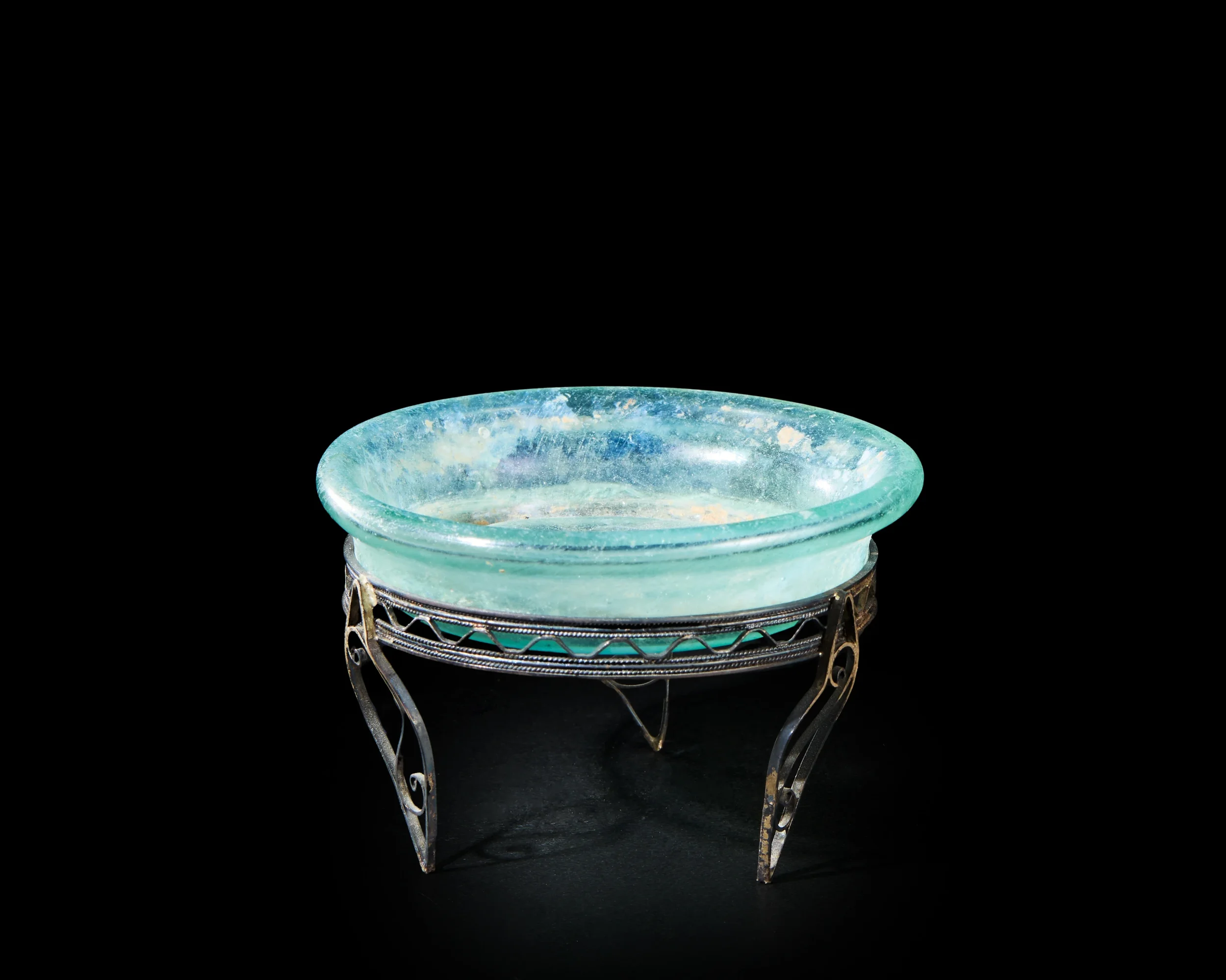A small, round glass dish with a slightly curved rim, resting on an ornate metal stand with curved legs, against a black background.