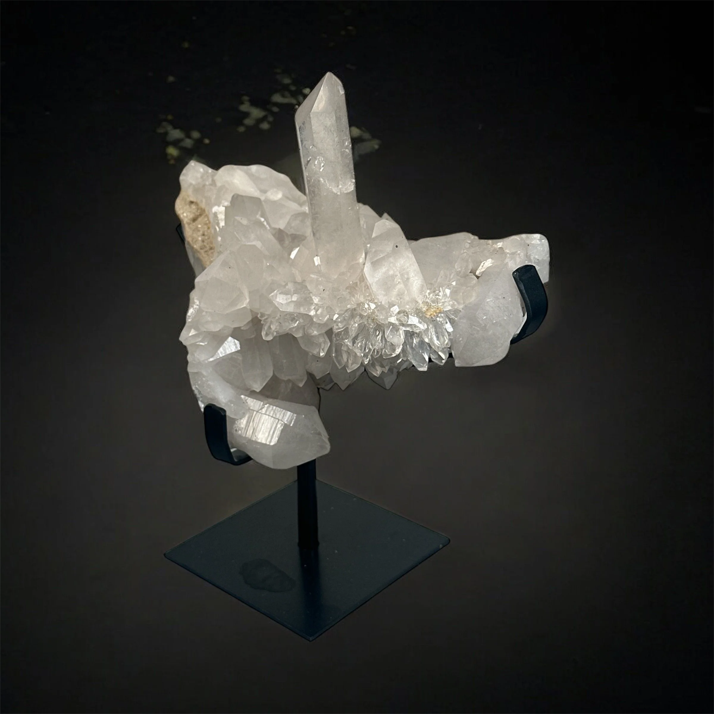 A crystal formation display with a large central clear crystal surrounded by smaller crystals, mounted on a black stand against a black background.