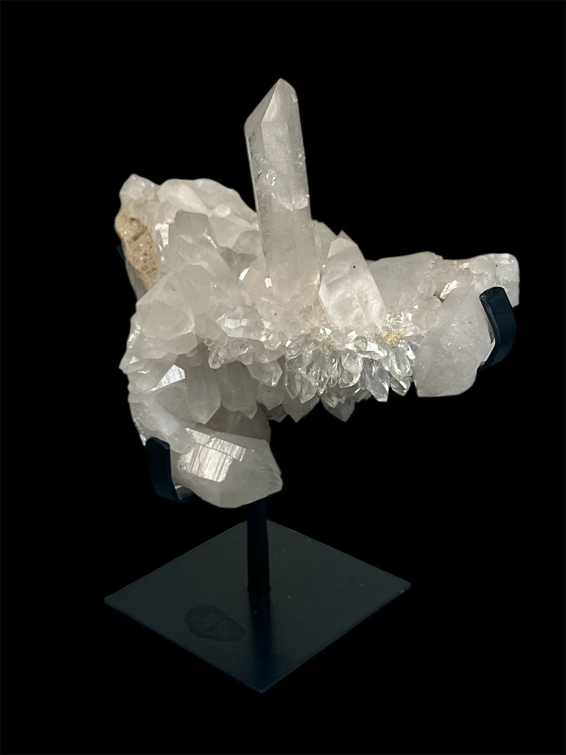 A crystal cluster with a tall, pointed quartz crystal surrounded by smaller clear and white crystals, displayed on a black stand against black background.