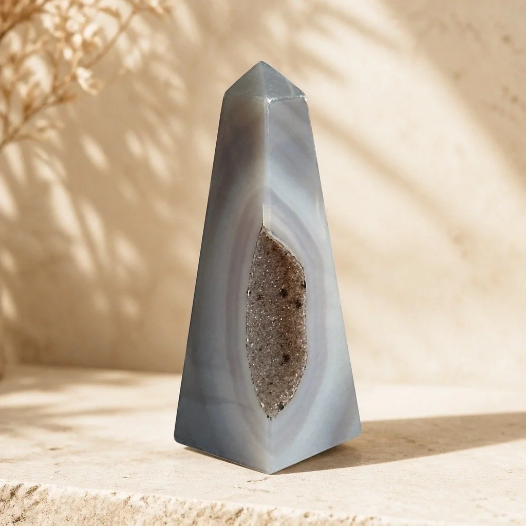A polished gray stone pyramid with a central geode cavity, placed on a textured surface with a blurred background and soft shadows.