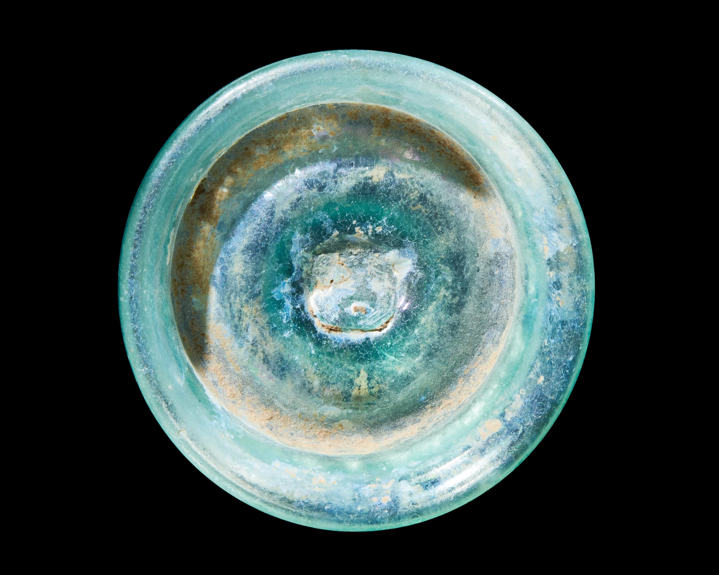 Close-up of an ancient, weathered, circular turquoise glass object with a central depression, possibly a artifact or decorative piece, against a black background.