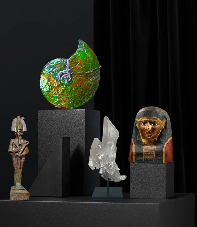 A display of Egyptian artifacts includes a bronze Osiris, a large colorful ammonite fossil, a clear quartz crystal, and a mummy mask