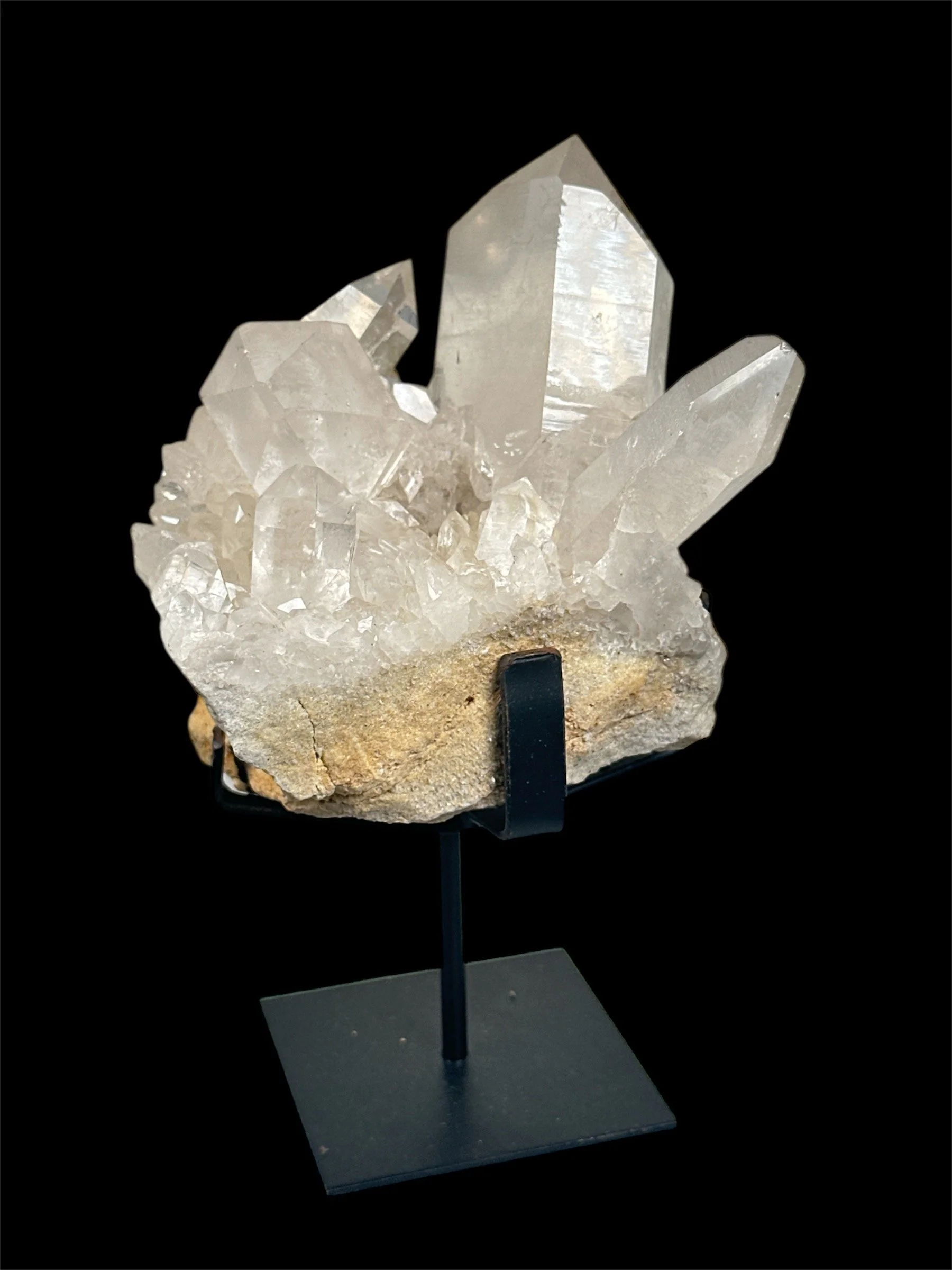 A crystal cluster with multiple large, clear quartz points on a rough base, displayed on a black stand against a black background.