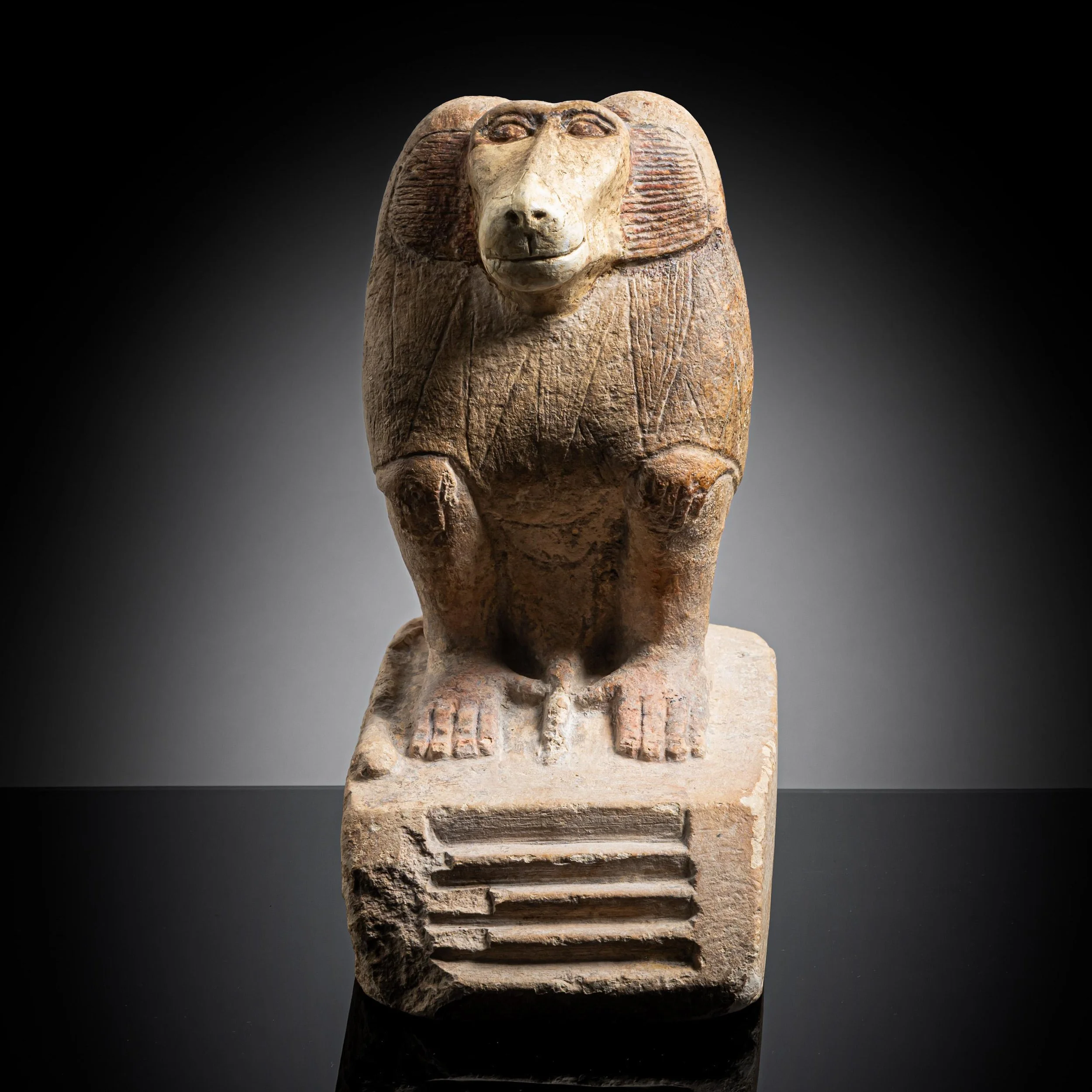 Ancient carved stone sculpture of a baboon with detailed mane and facial features, mounted on a base, against a dark background.