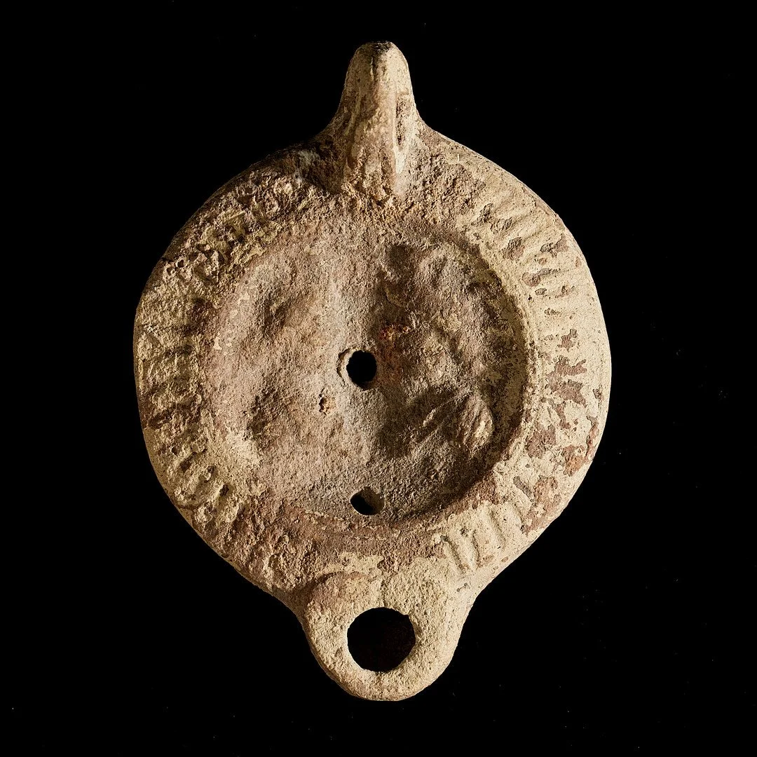 Ancient clay oil lamp with a round body, three small openings, and a pointed handle, on a black background.