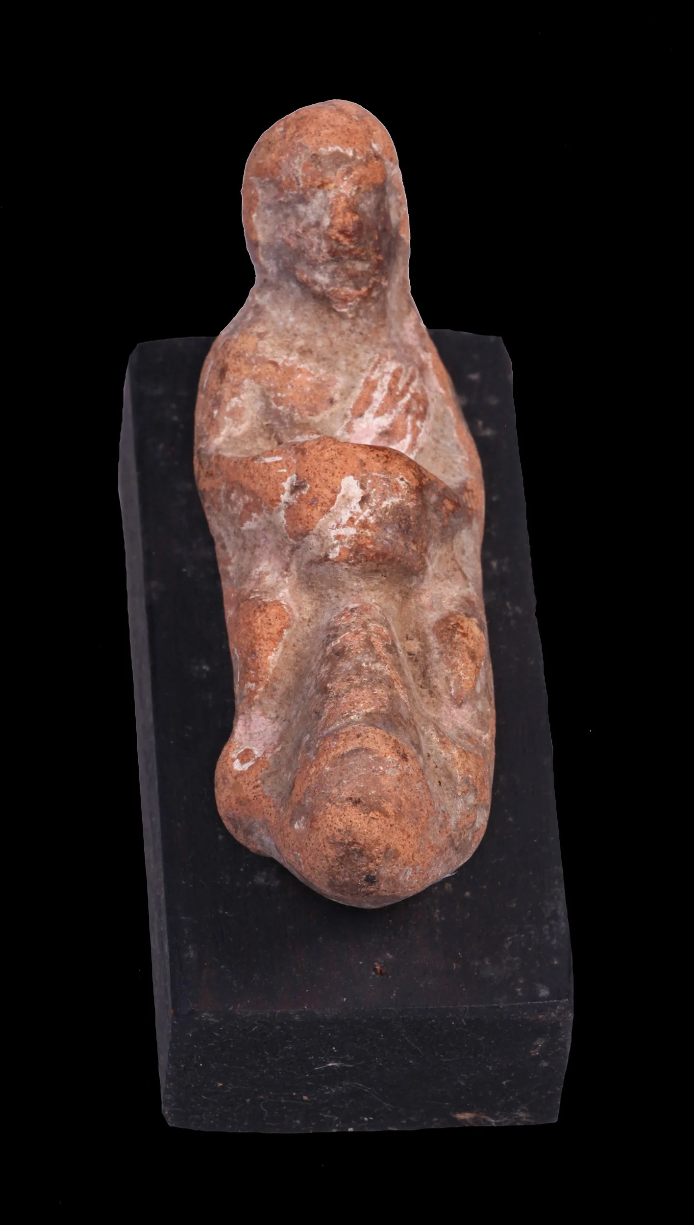 Ancient terracotta figurine of a woman with a hand placed on her chest, displayed on a black pedestal.