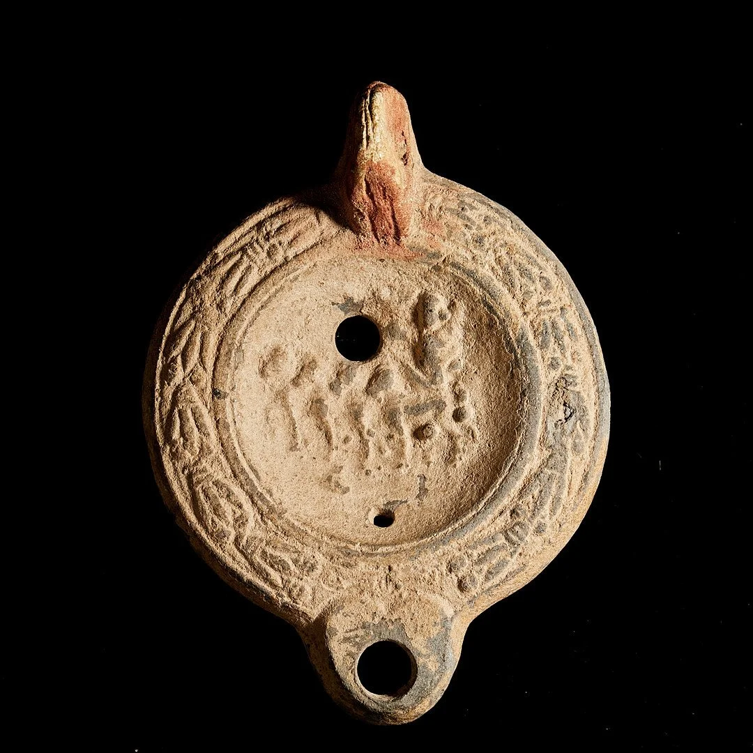 Ancient ceramic or clay oil lamp or pendant with circular design and engraved symbols, including a central motif and a raised handle, against a black background.