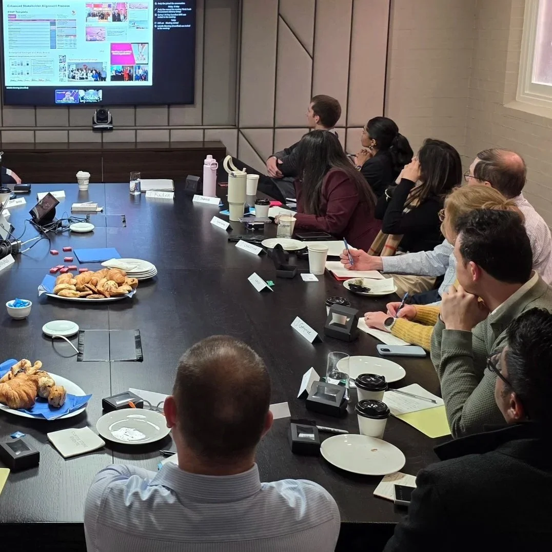 People attending a meeting in a conference room, watching a presentation on a large screen, with food and drinks on the table.