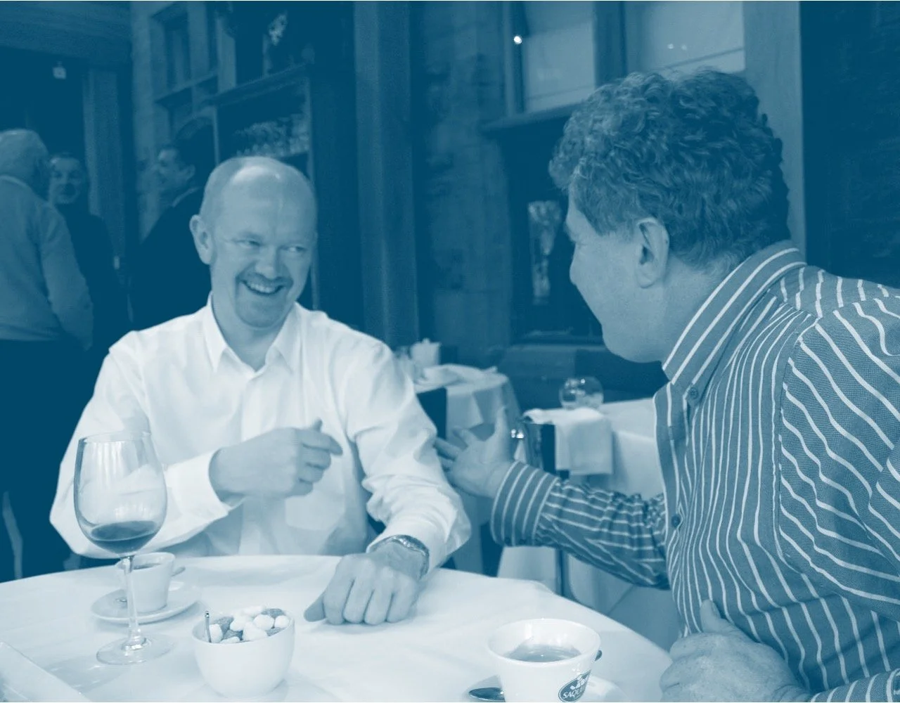 Two men having a lively conversation at a restaurant table, one wearing a white shirt and the other in a striped shirt, with glasses of wine and tea on the table.