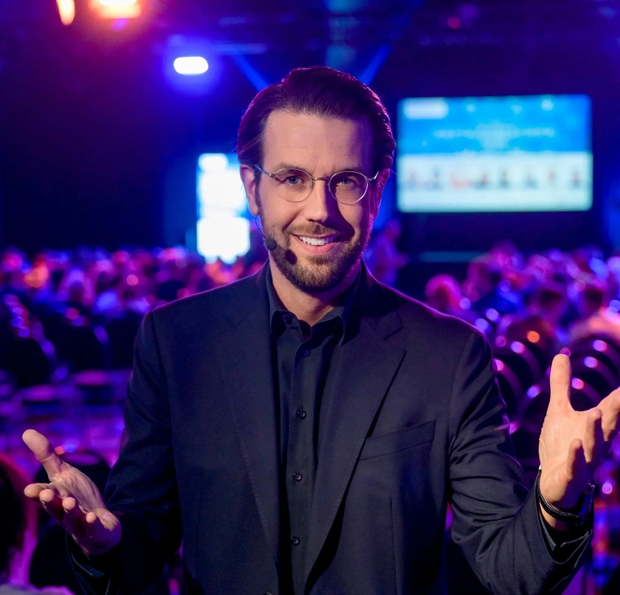 Ein Mann mit Brille, dunklem Anzug und Headset auf einer Bühne bei einer Veranstaltung, umgeben von einem Publikum im dunklen Raum mit blauer und lila Beleuchtung.