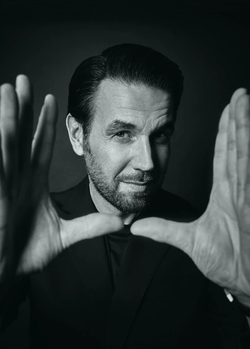Schwarz-weiß-Foto eines Mannes mit dunklem Haar und Bart, der die Hände in einer Rahmenposition hält, blickt direkt in die Kamera.