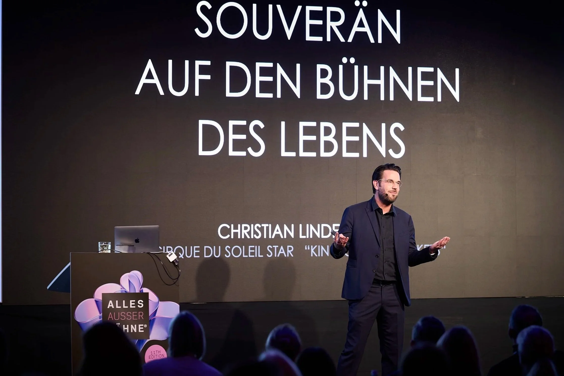 Mann in dunklem Anzug spricht vor Publikum bei Vortrag oder Präsentation, großes Bildschirm im Hintergrund zeigt den Titel 'Souverän auf den Bühnen des Lebens' und den Namen 'Christian Linde'.