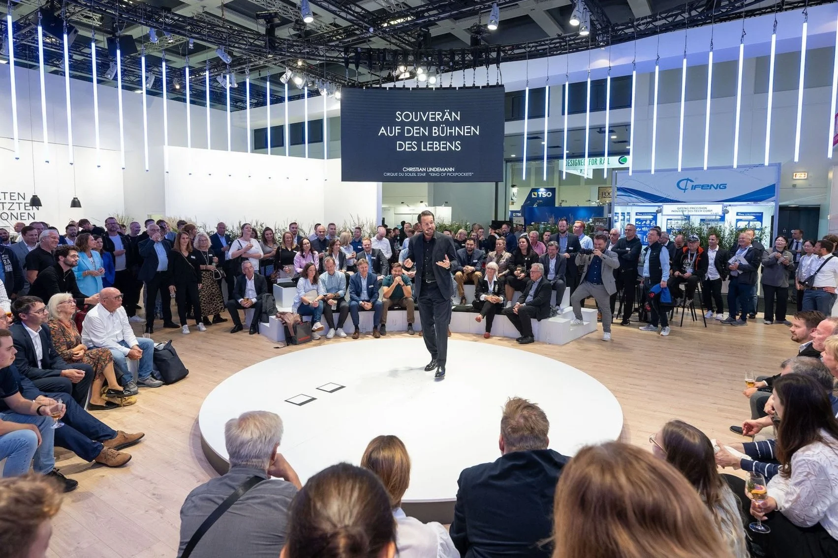 Ein Mann hält eine Rede oder führt eine Präsentation vor einer großen, sitzenden und stehenden Zuschauergruppe auf einer runden Bühne in einer modernen Ausstellungshalle.