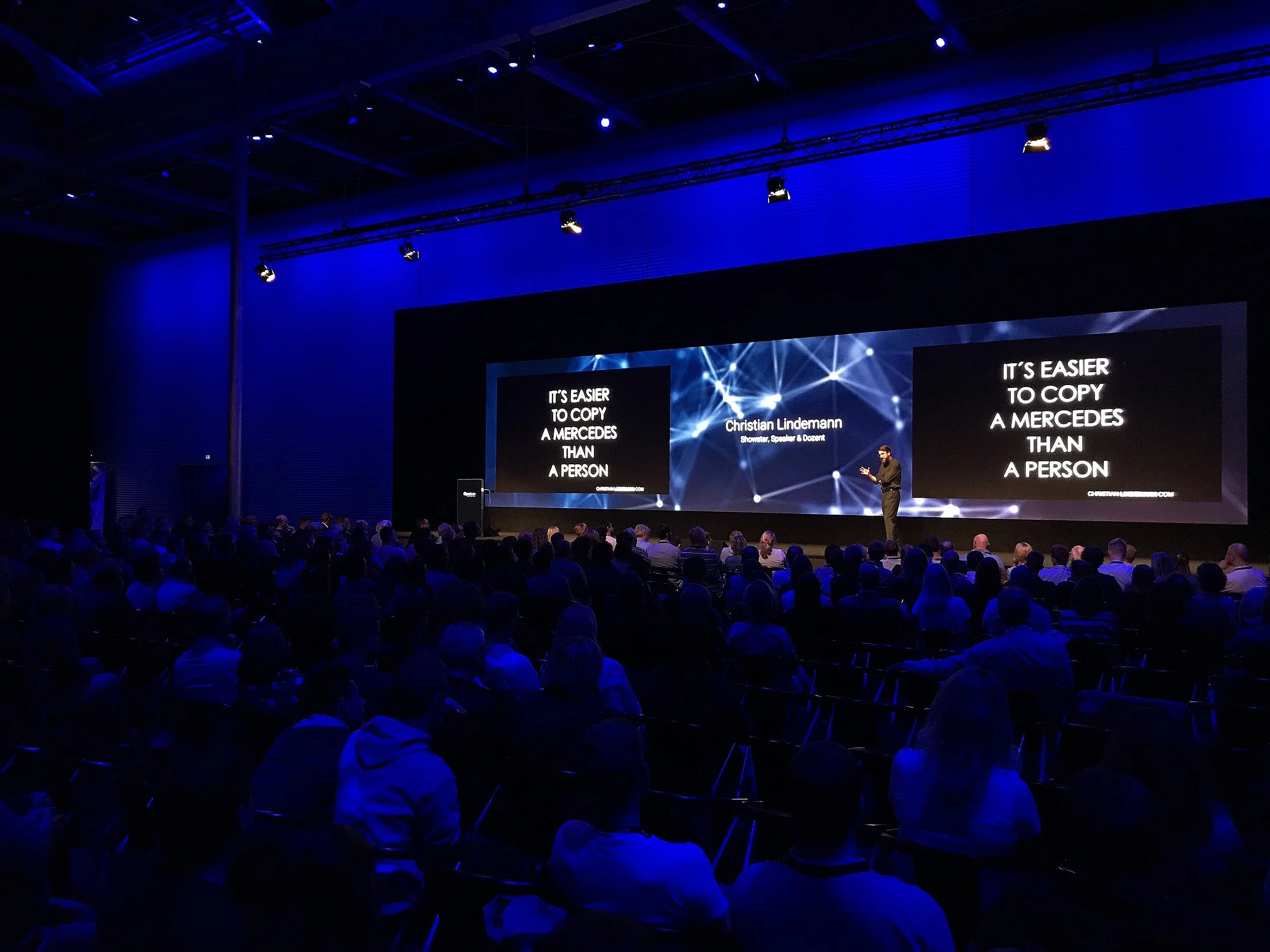 Ein dunkler Konzertsaal mit blauer Beleuchtung, voll mit Menschen, die einem Vortrag auf der Bühne folgen. Auf der großen Leinwand sind die Worte: 'Es ist einfacher, einen Mercedes zu kopieren als eine Person.' Im Hintergrund ist ein Sprecher, Christ