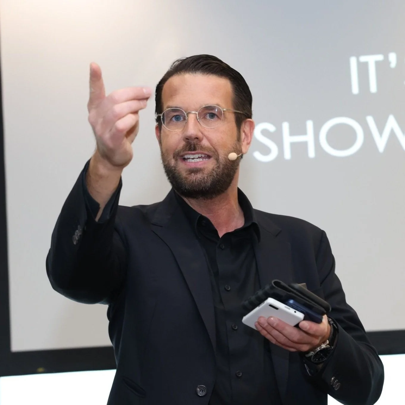 Ein Mann mit Brille und Bart hält ein Mikrofon in der Hand und zeigt mit der anderen Hand nach vorne. Im Hintergrund ist ein Bildschirm mit dem Text 'IT'S SHOW'.