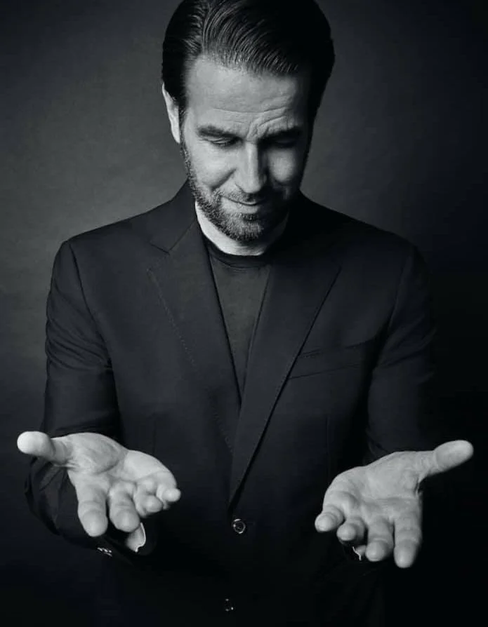 Schwarz-weiß-Foto eines Mannes in dunklem Anzug, der mit gesenktem Blick die Handflächen nach oben hält, wirkt nachdenklich.