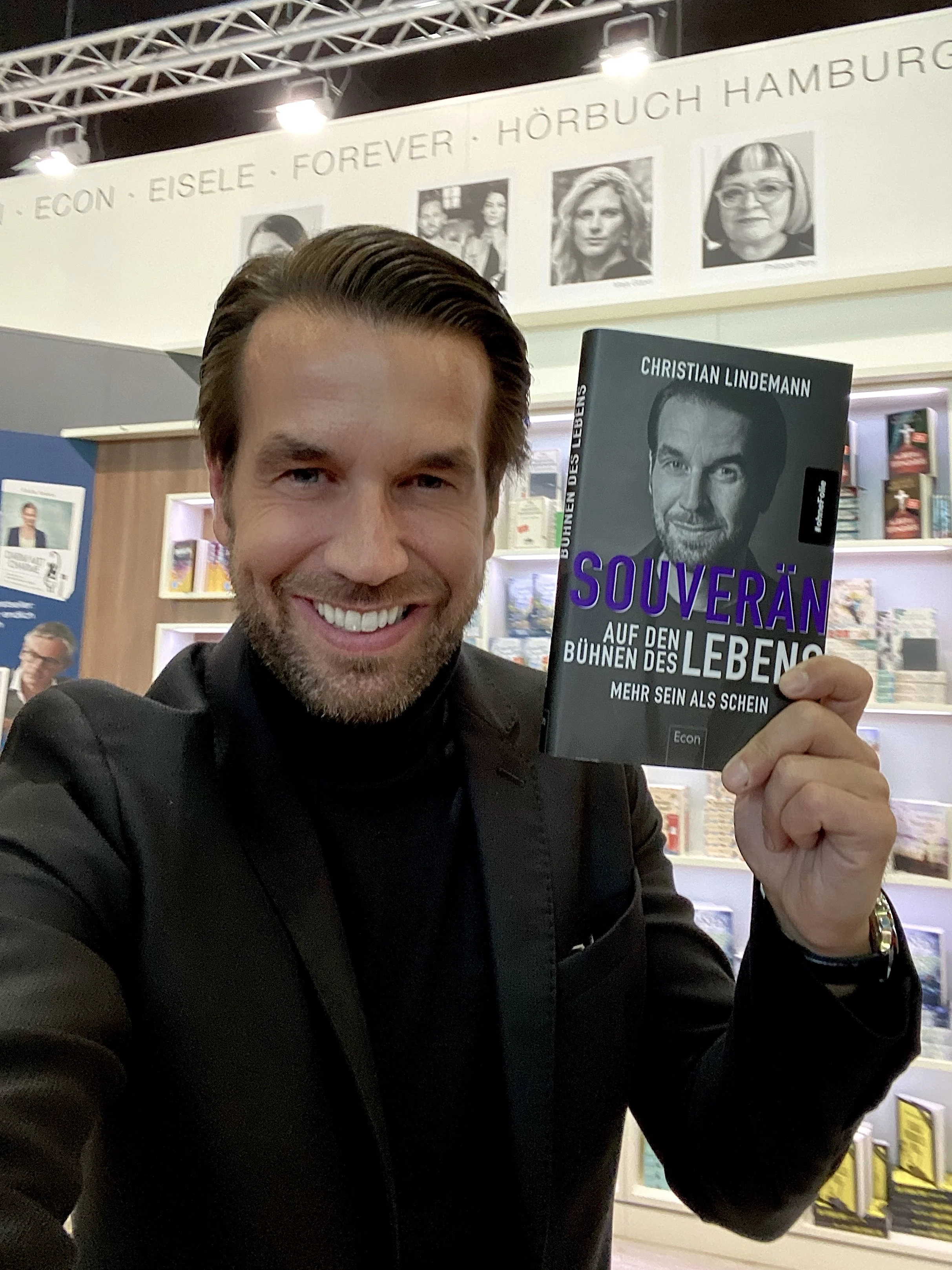 Ein lachender Mann hält ein Buch mit dem Titel 'Souverän auf den Bühnen des Lebens' bei einer Buchmesse. Im Hintergrund sind Bücherregale und Plakate zu sehen.