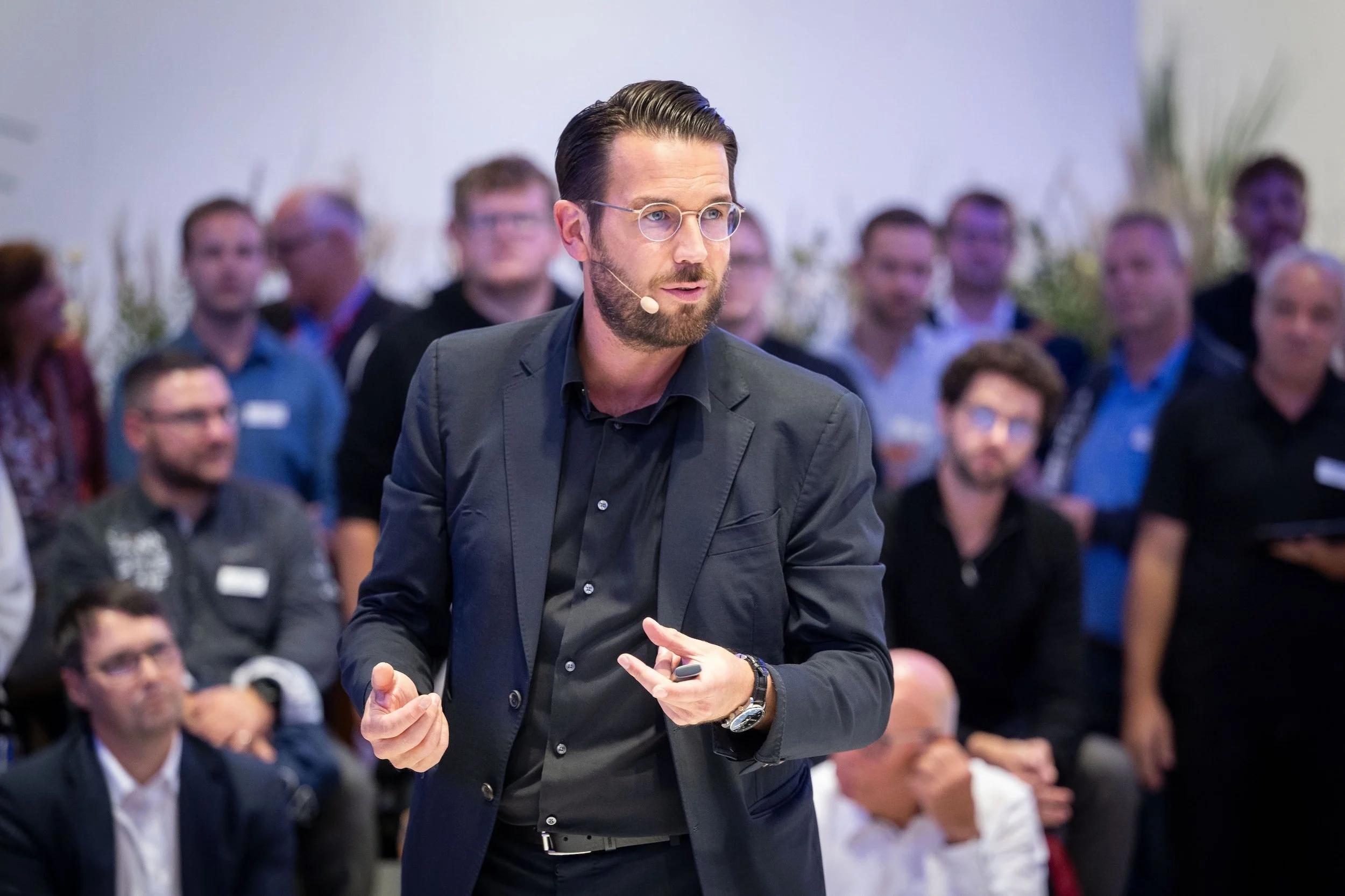Ein Mann mit Brille und dunklem Anzug hält eine Präsentation vor einem Publikum bei einer Konferenz.