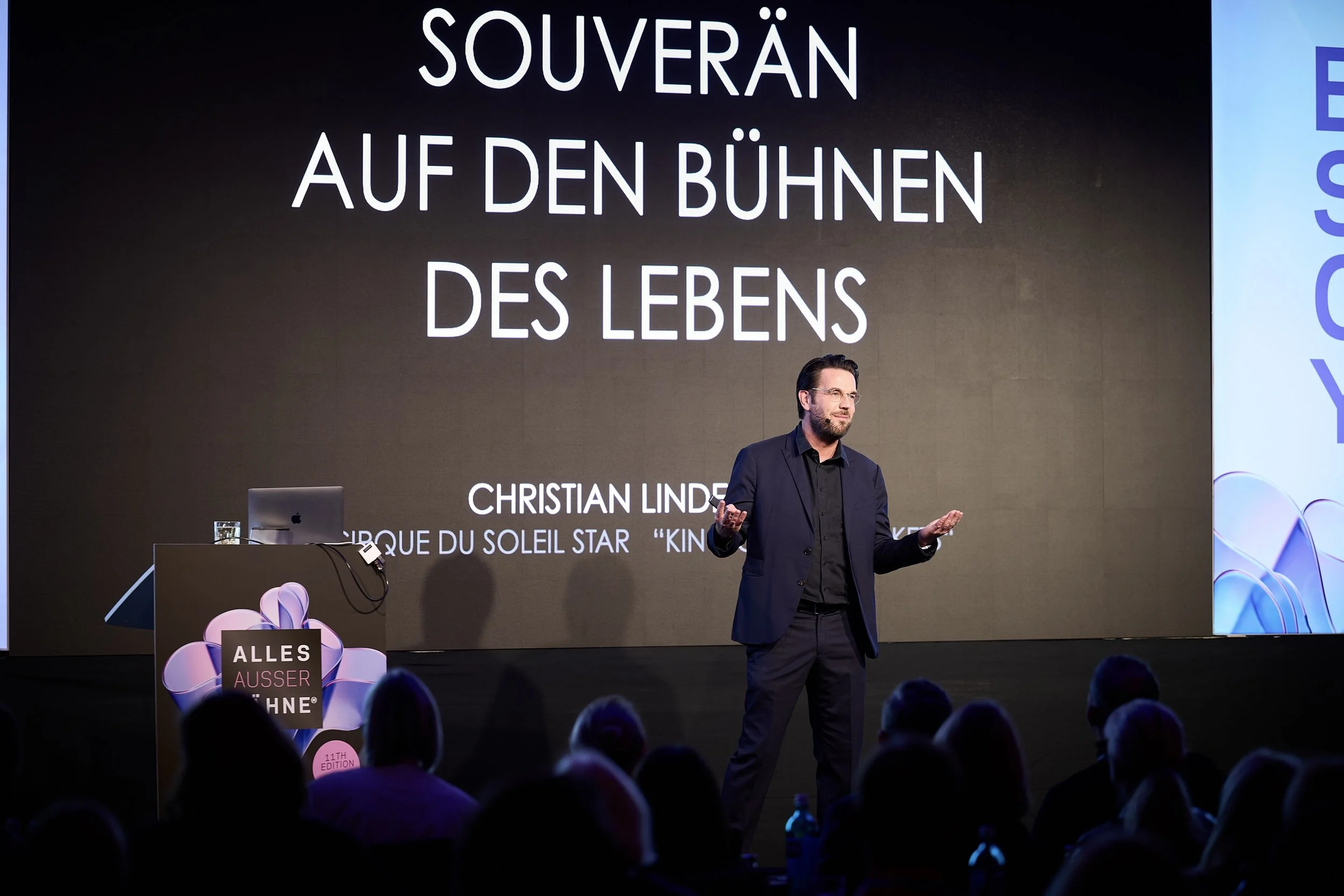 Mann gestikuliert auf einer Bühne vor einer großen Leinwand mit dem Text »Souverän auf den Bühnen des Lebens«. Im Vordergrund sitzen Zuschauer, und es gibt eine Tafel mit einem Blumenmotiv und dem Schriftzug »Alles außer schön«.