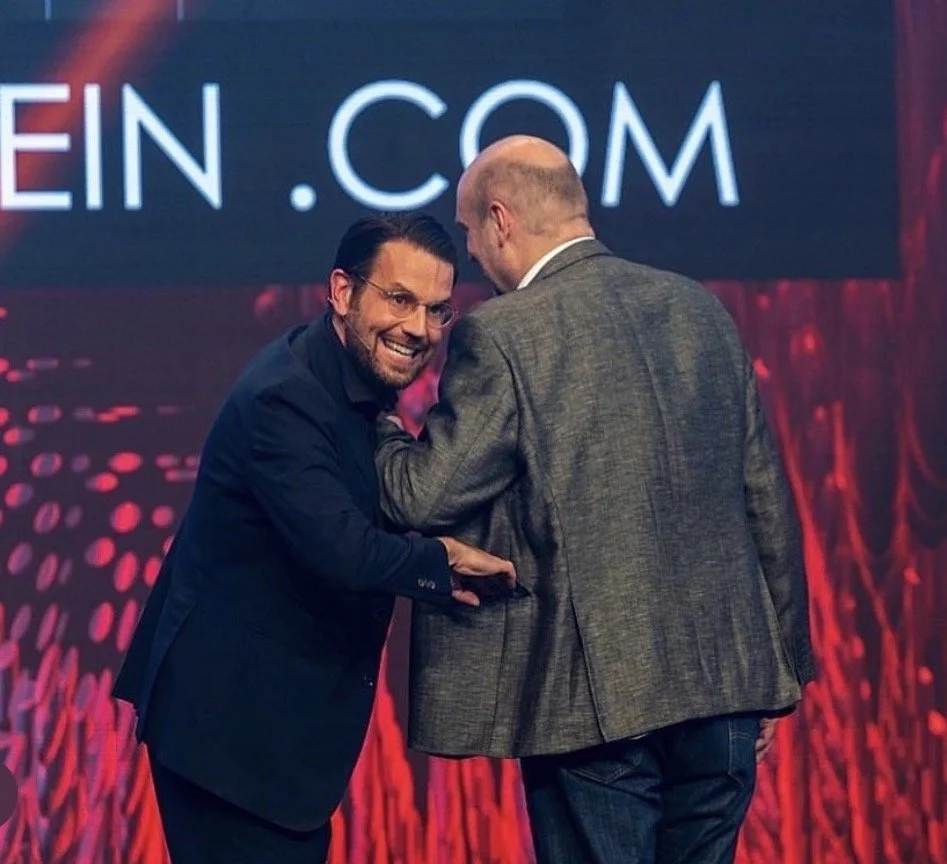 Zwei Männer auf einer Bühne unterhalten sich und lachen. Der Hintergrund zeigt ein großes digitales Display mit Text und roten Lichtern.