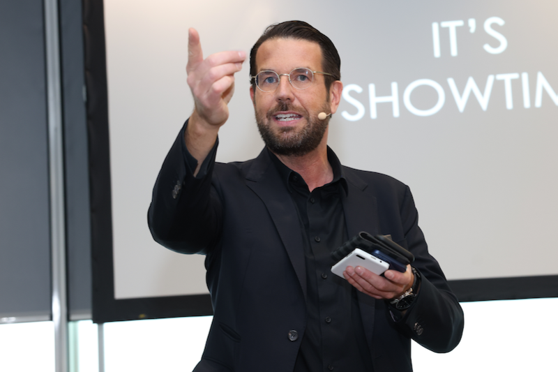 Mann hält ein Mikrofon und ein Smartphone, spricht vor Publikum, hinter ihm eine Leinwand mit der Aufschrift "IT'S SHOWTIME".