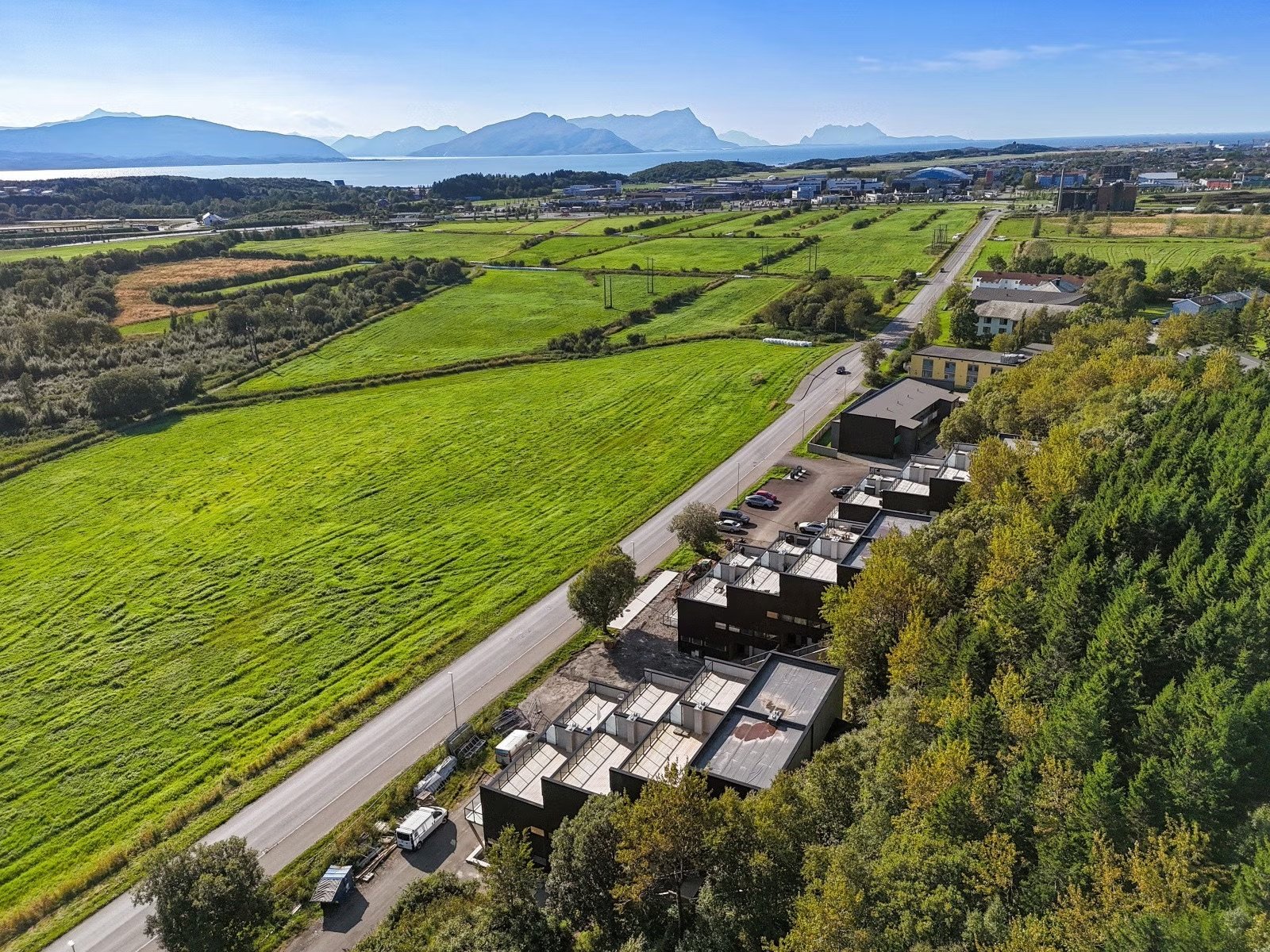 Aerial view of green fields, a road, and residential buildings with trees, mountains, and water in the distance on a sunny day.