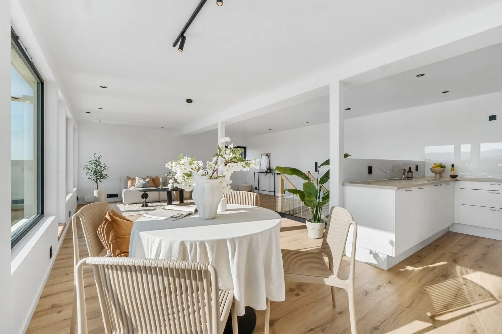 Bright open-plan living and dining area with white walls, wooden floors, large windows, a dining table with a white tablecloth, woven chairs, a vase of white flowers, a green potted plant, and a kitchen with white cabinets and a countertop.