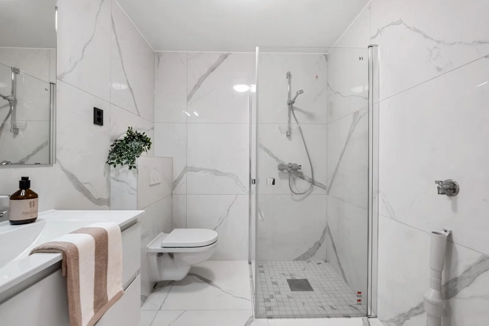 Modern bathroom with white marble walls, a glass shower enclosure, a wall-mounted toilet, a white vanity with a towel, a mirror, a plant, and toiletries.