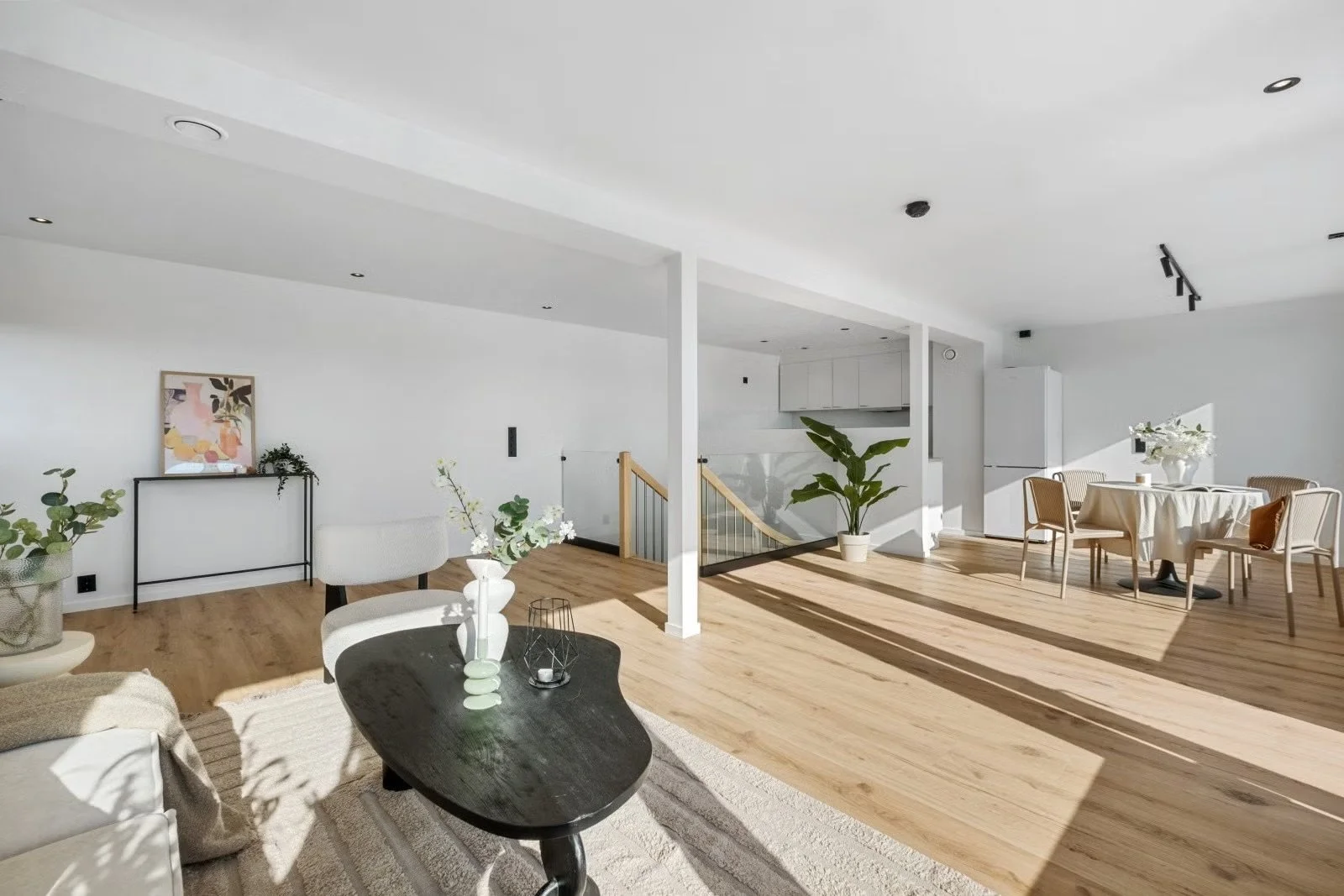 Modern open-concept living and dining area with white walls, wooden floors, and minimalistic furniture. There are plants, a black coffee table, a dining table with chairs, and a kitchen area in the background.