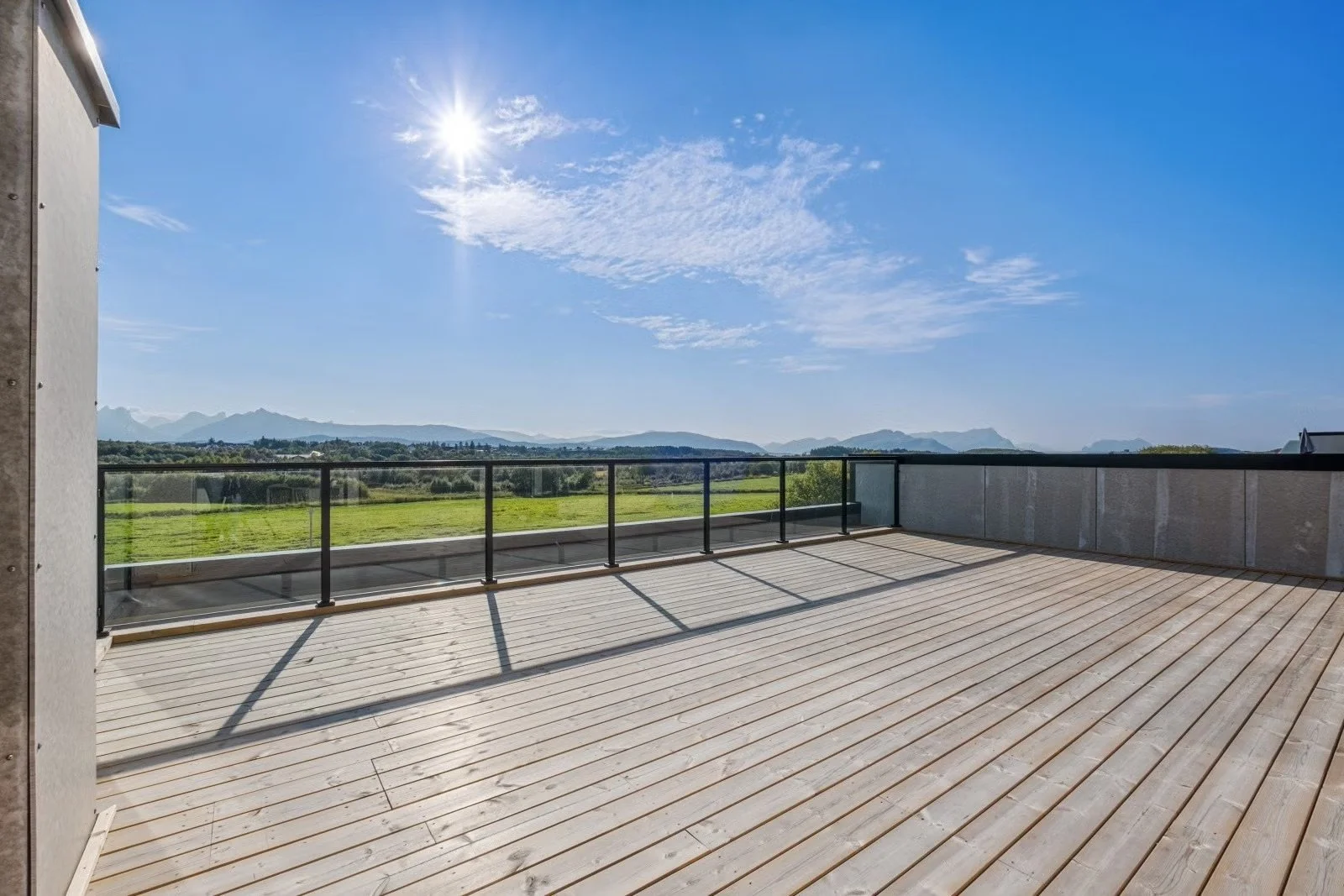 Empty balcony with wooden flooring, glass railing, and panoramic view of green fields, mountains, and a clear blue sky with the sun shining brightly.