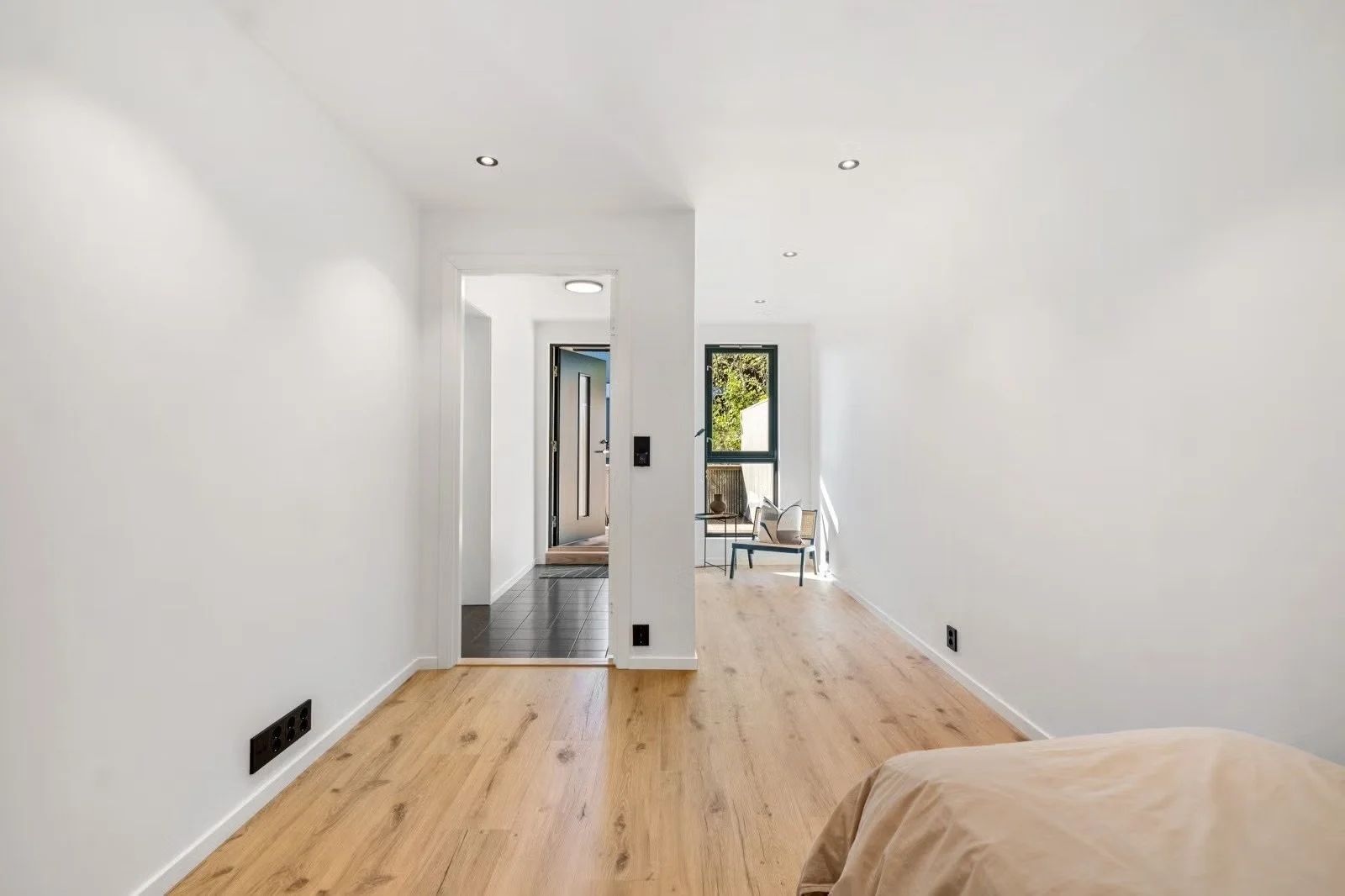 Interior of a modern, minimalist apartment with white walls and wooden flooring, showing a hallway leading to a small seating area near a window with a chair and a small table.