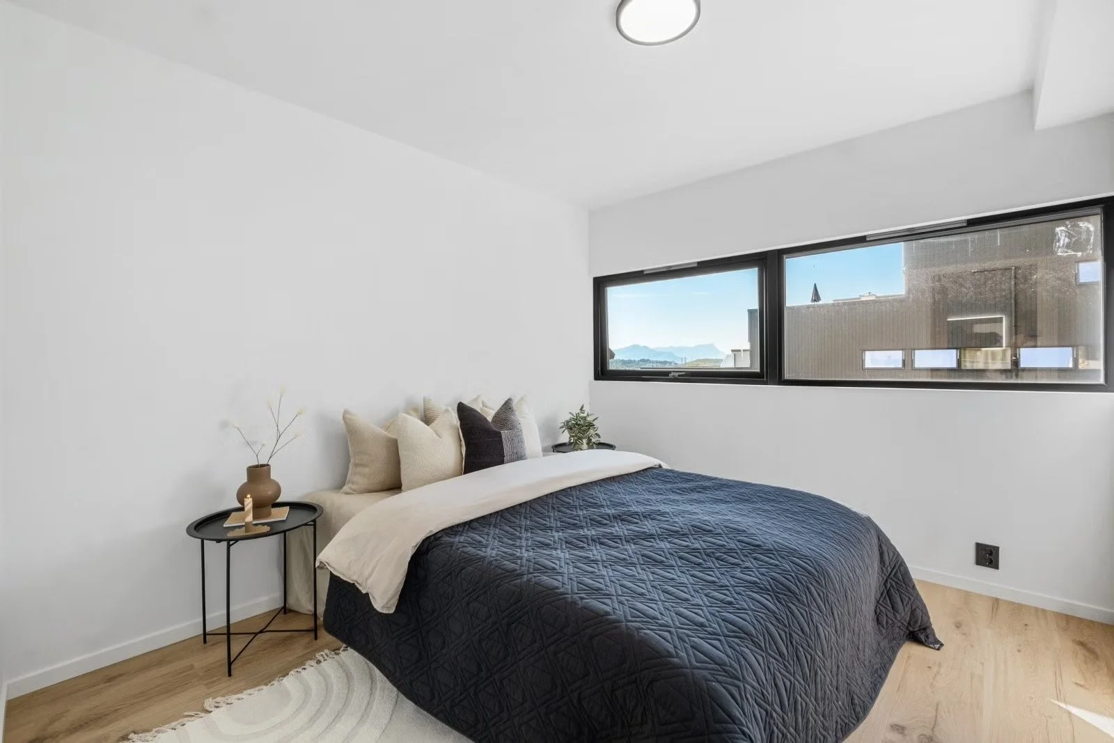 Minimalist bedroom with a bed featuring a black quilt and beige and black pillows, a side table with a vase and candle, and large windows showing outdoor scenery.