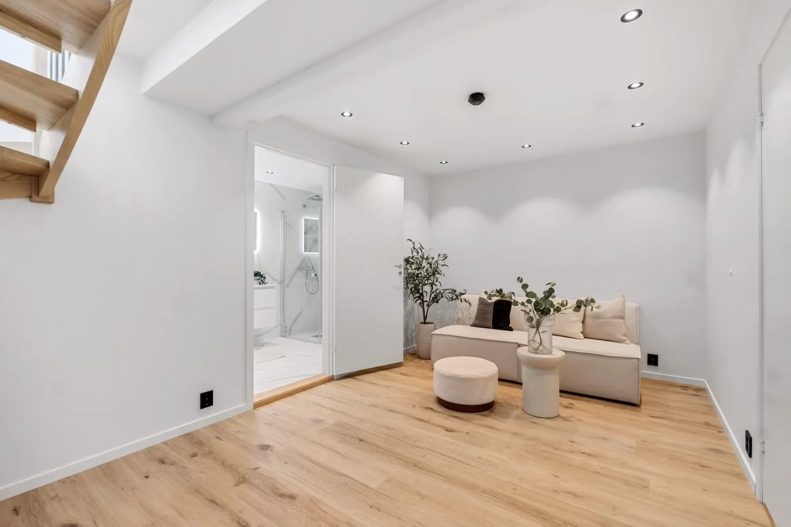 Minimalist living room with white walls, wooden flooring, and a small sofa with cushions, decorated with potted plants, under recessed ceiling lights, with a view into a bathroom with a shower.