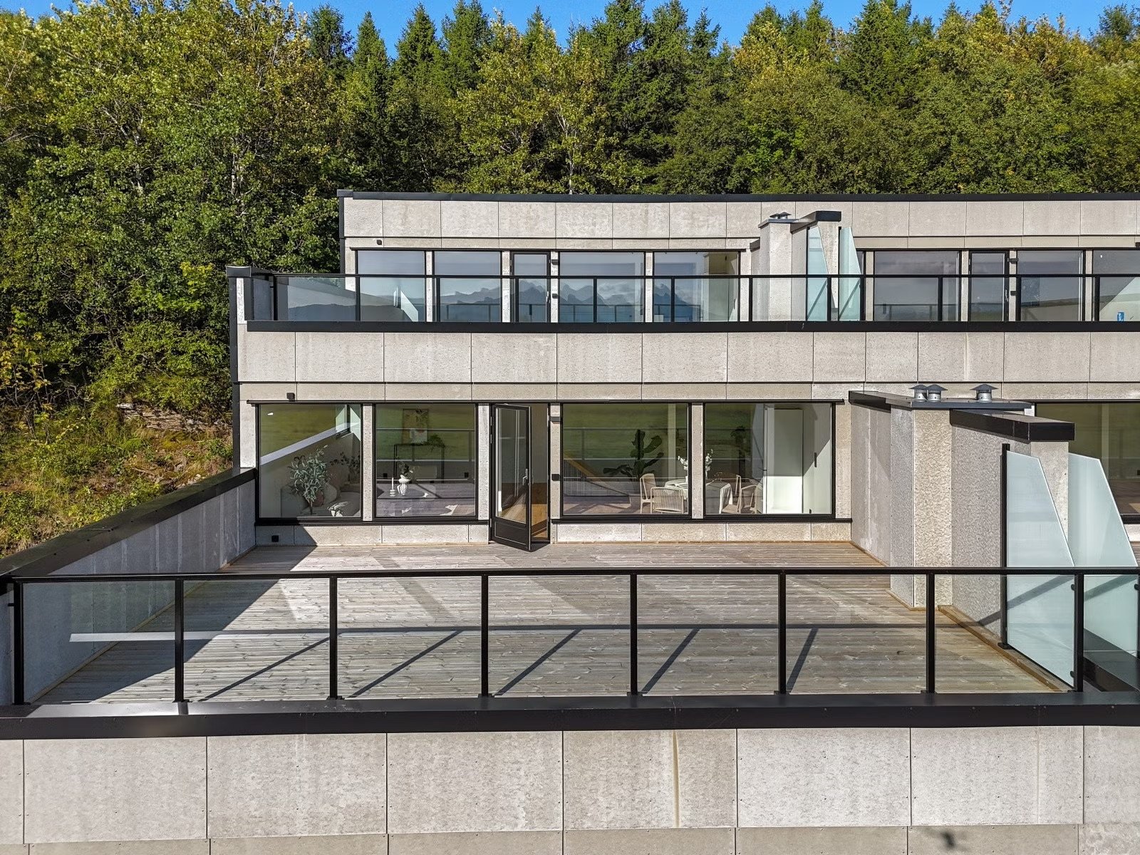 Modern multi-story building with large glass windows and a spacious outdoor balcony or terrace, surrounded by lush green trees.