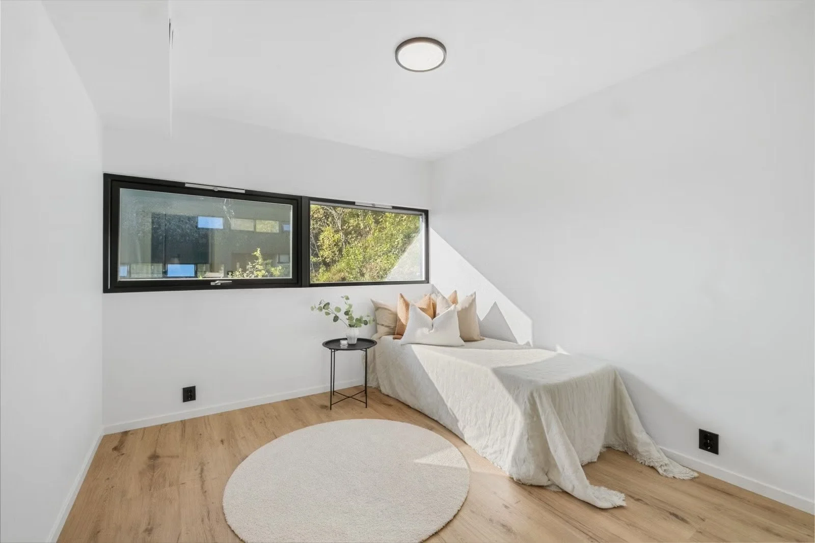 Minimalist bedroom with a bed covered in a white quilt and pillows, a small black side table with a plant, a large window with a view of greenery, wooden floor, and off-white walls.