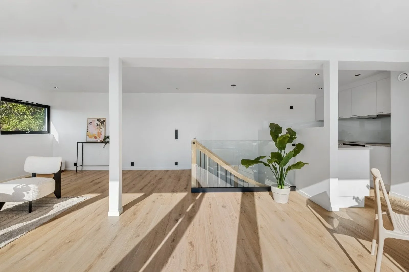 Modern, minimalist living space with light wood flooring, white walls, a large window with greenery outside, a potted plant, and minimalist furniture including a white chair and a small side table with artwork.