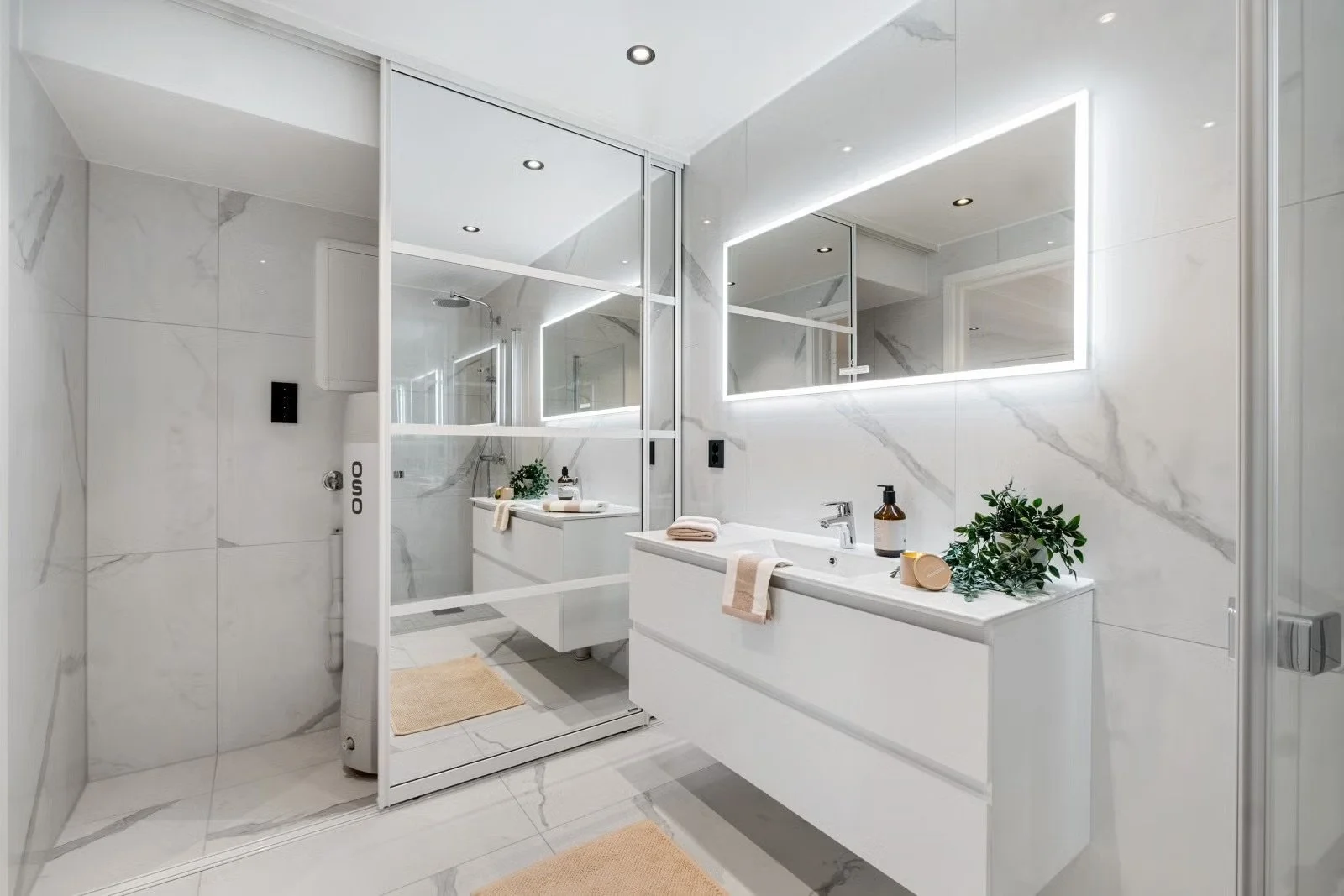 Modern bathroom with marble walls, large mirror with LED lighting, white vanity with sink, plants, towels, and bathroom accessories. The shower area has a glass door and a rain shower head.
